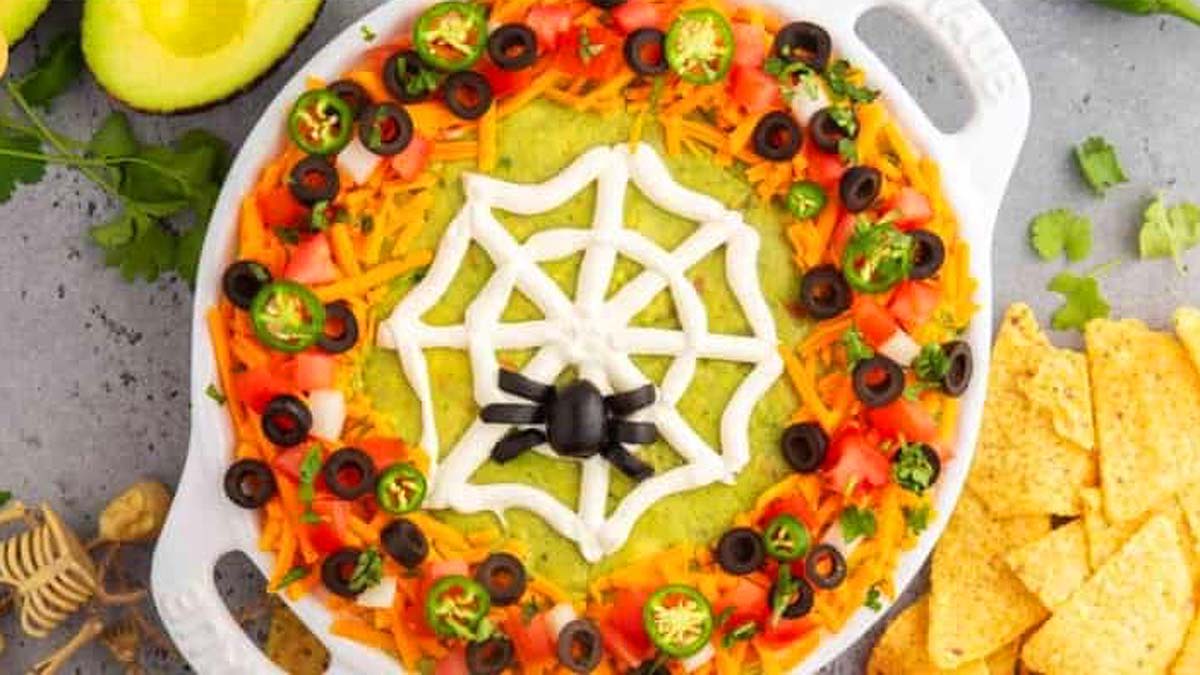 A dish of taco dip with a sour cream web and an olive spider.