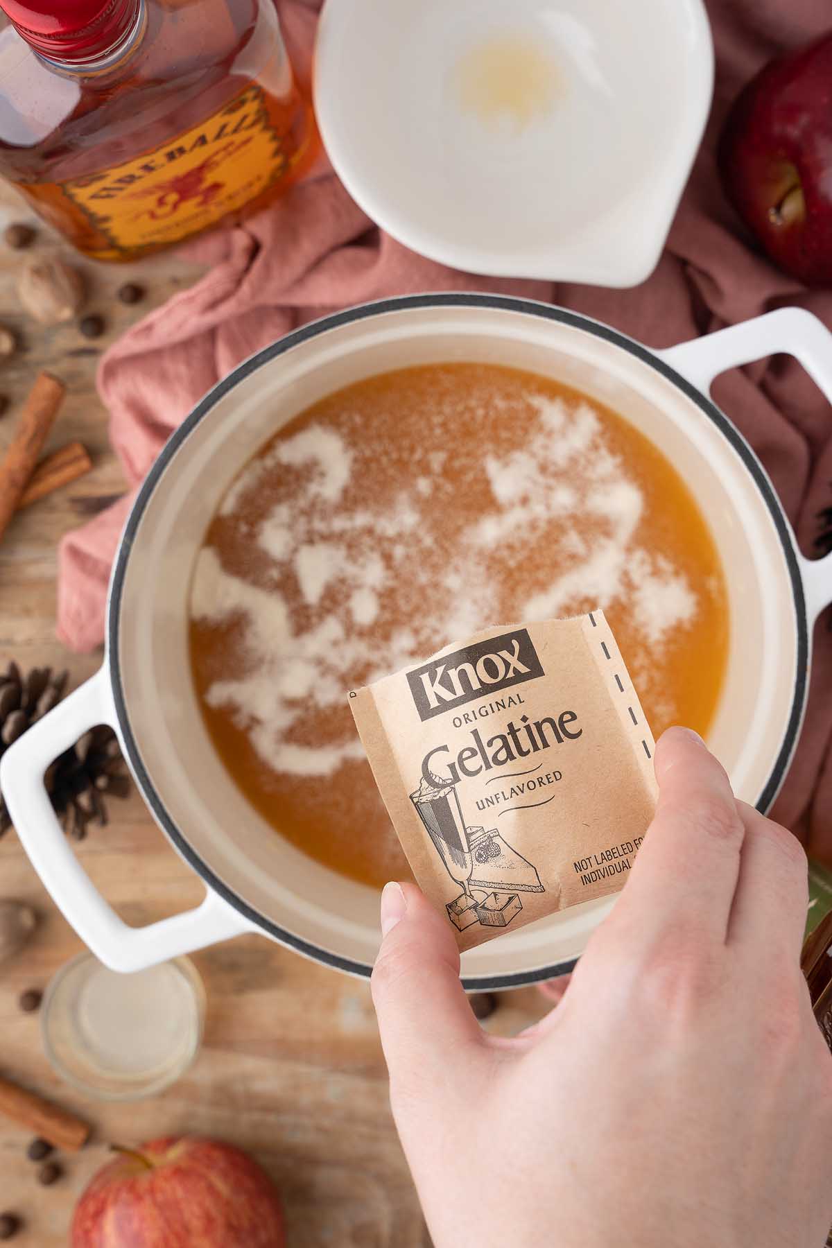 A hand holds a packet of unflavored gelatin over a pot of light brown liquid on a wooden table, perfect for making apple pie jello shots.