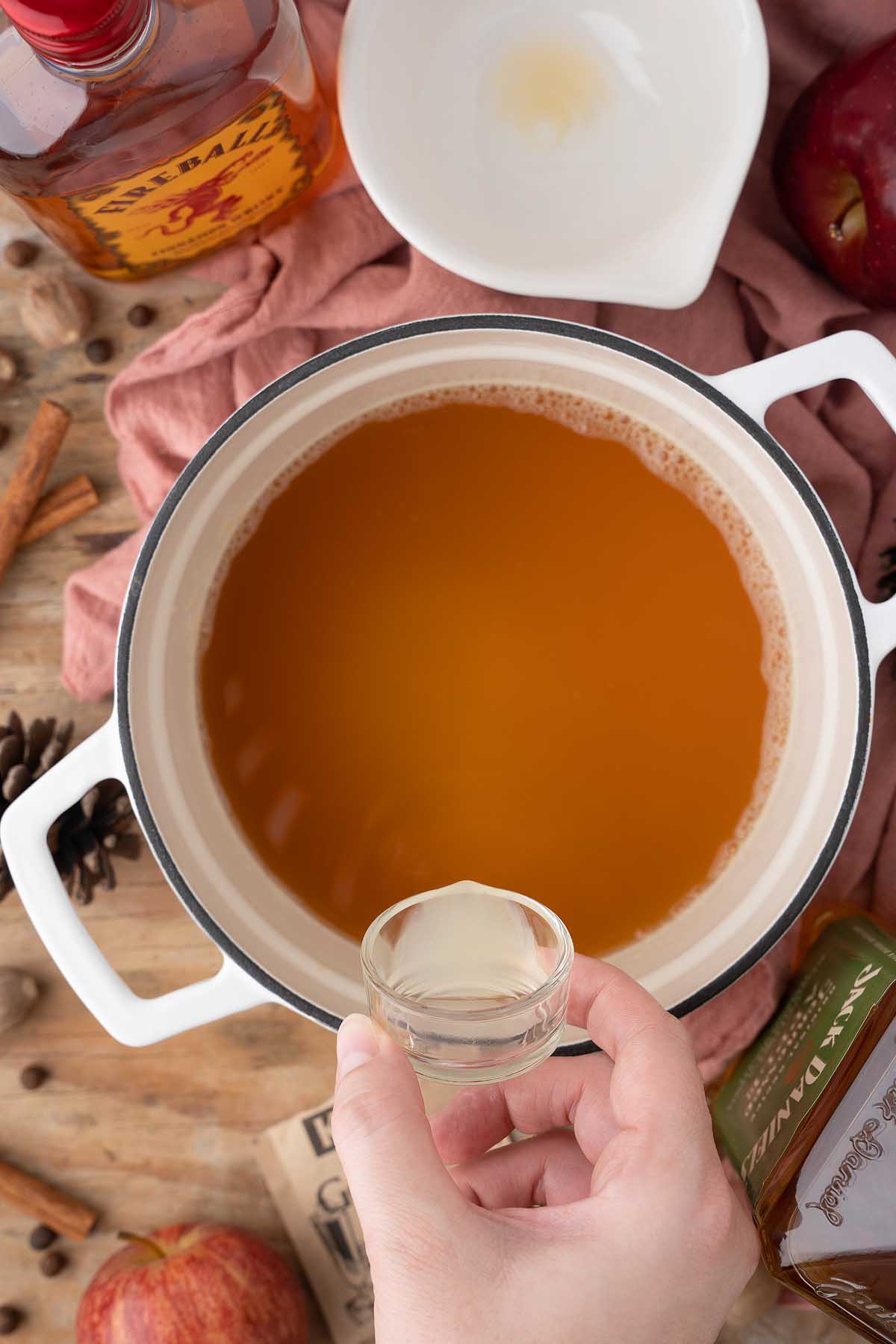 Hand holding a shot glass over a pot of spiced apple cider, surrounded by whiskey, apples, cinnamon sticks, and festive apple pie jello shots.
