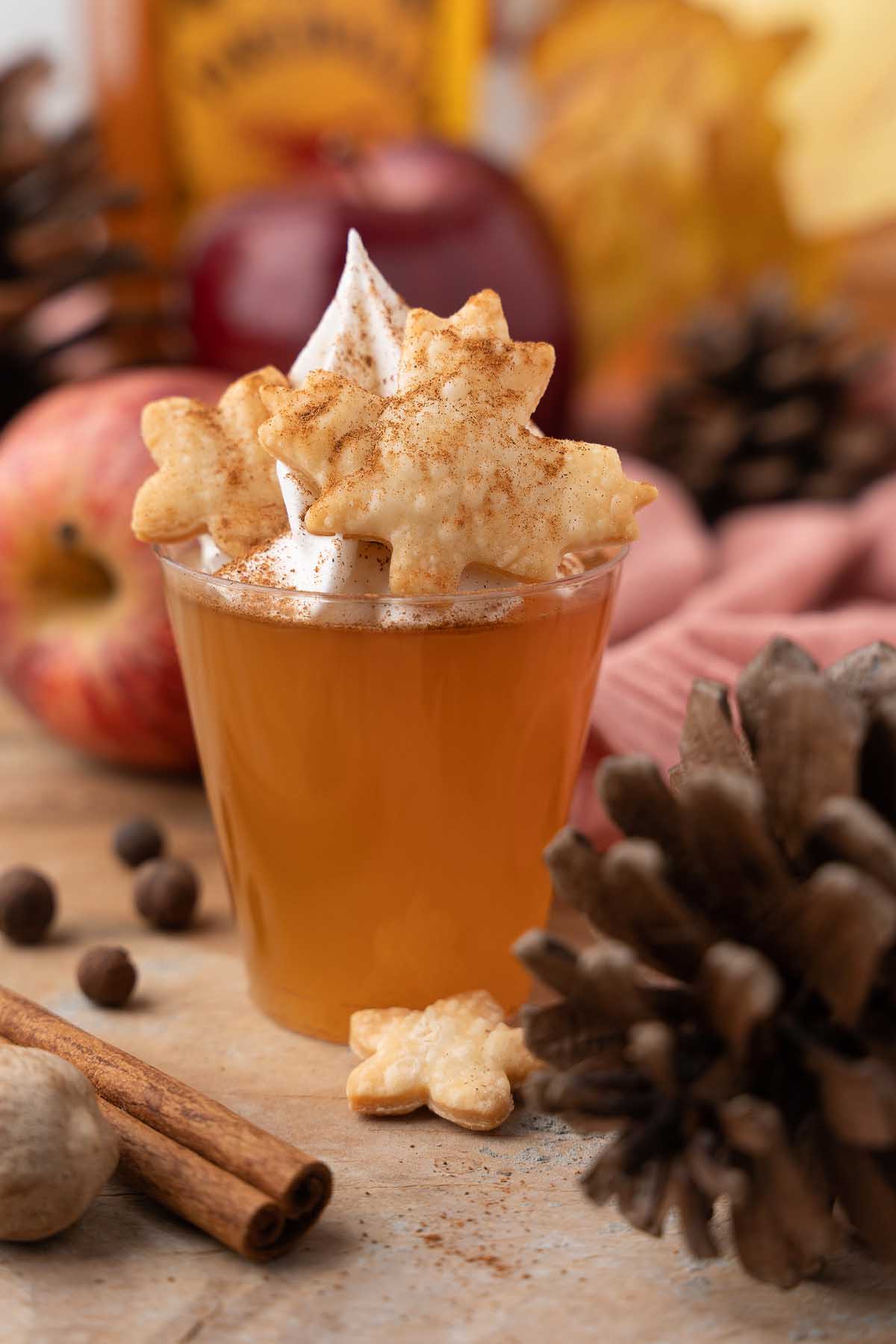 A glass of apple cider topped with whipped cream and leaf-shaped cookies, surrounded by autumn decor, evokes the cozy spirit of fall—perfect alongside festive apple pie jello shots.