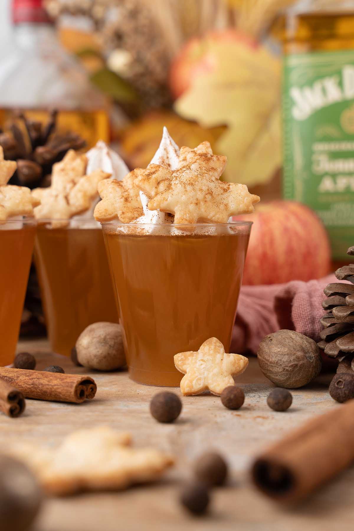 A spiced apple cocktail inspired by apple pie jello shots, topped with star-shaped pie crust and whipped cream, surrounded by fall spices.