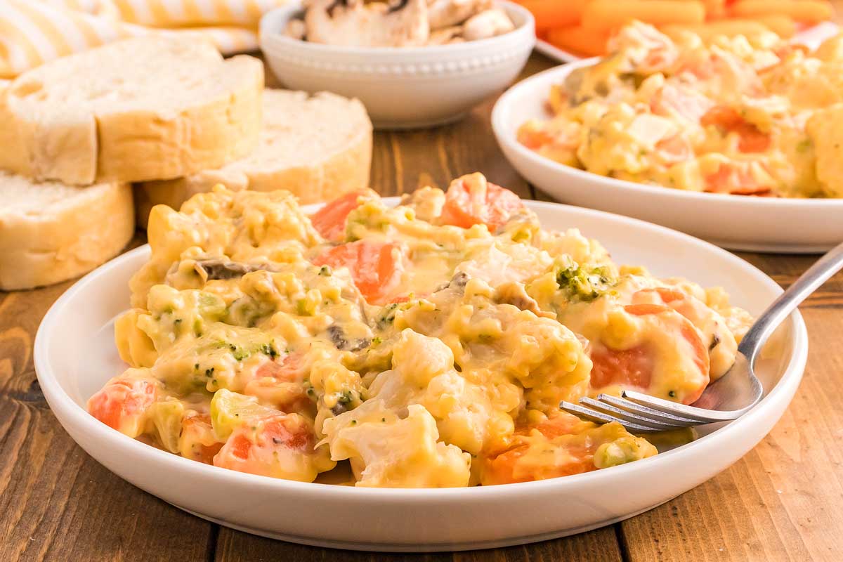 A bowl of creamy vegetable pasta featuring carrots, broccoli, and mushrooms. The dish is topped with a rich yellow sauce, and is accompanied by slices of bread and a small bowl of mushrooms in the background. A fork rests inside the bowl.