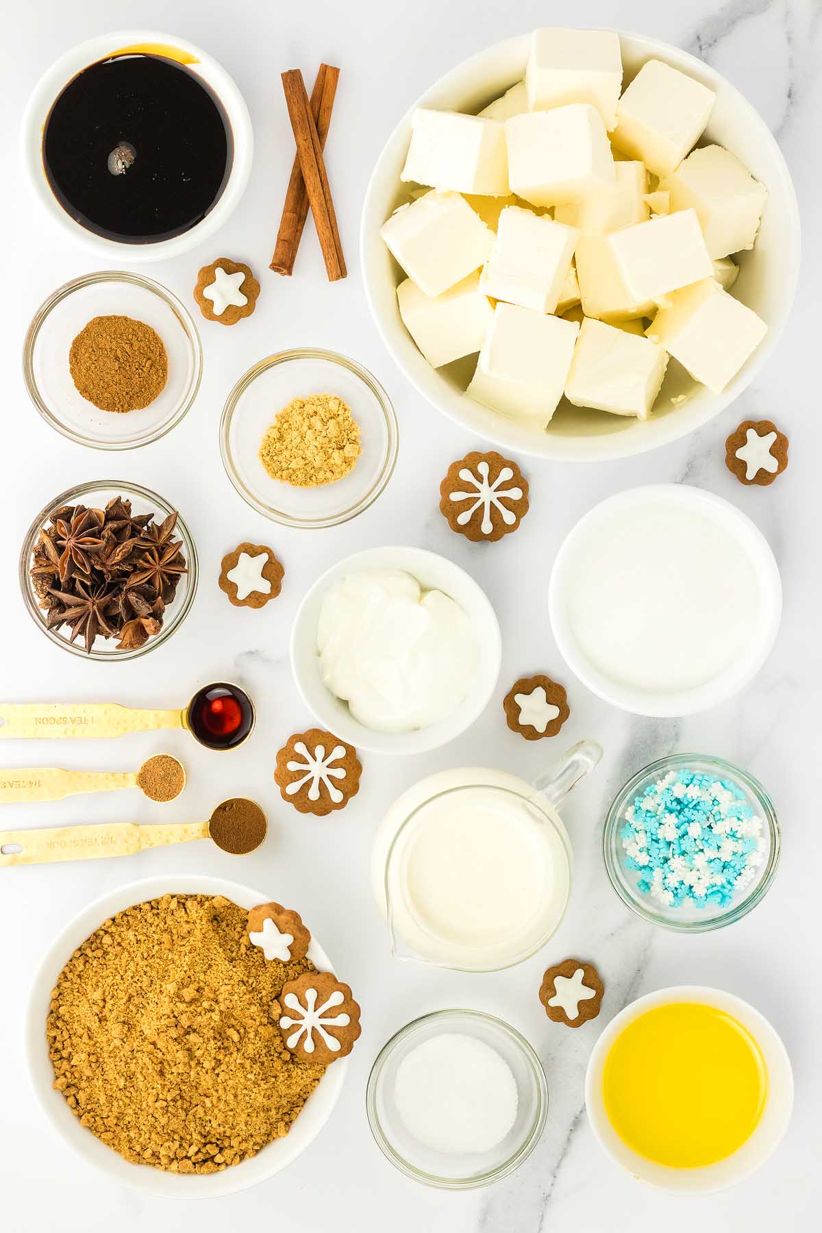 Baking ingredients in bowls and measuring spoons, including butter, cream, spices, sugar, and small decorated cookies—perfect for making gingerbread cheesecake jars.