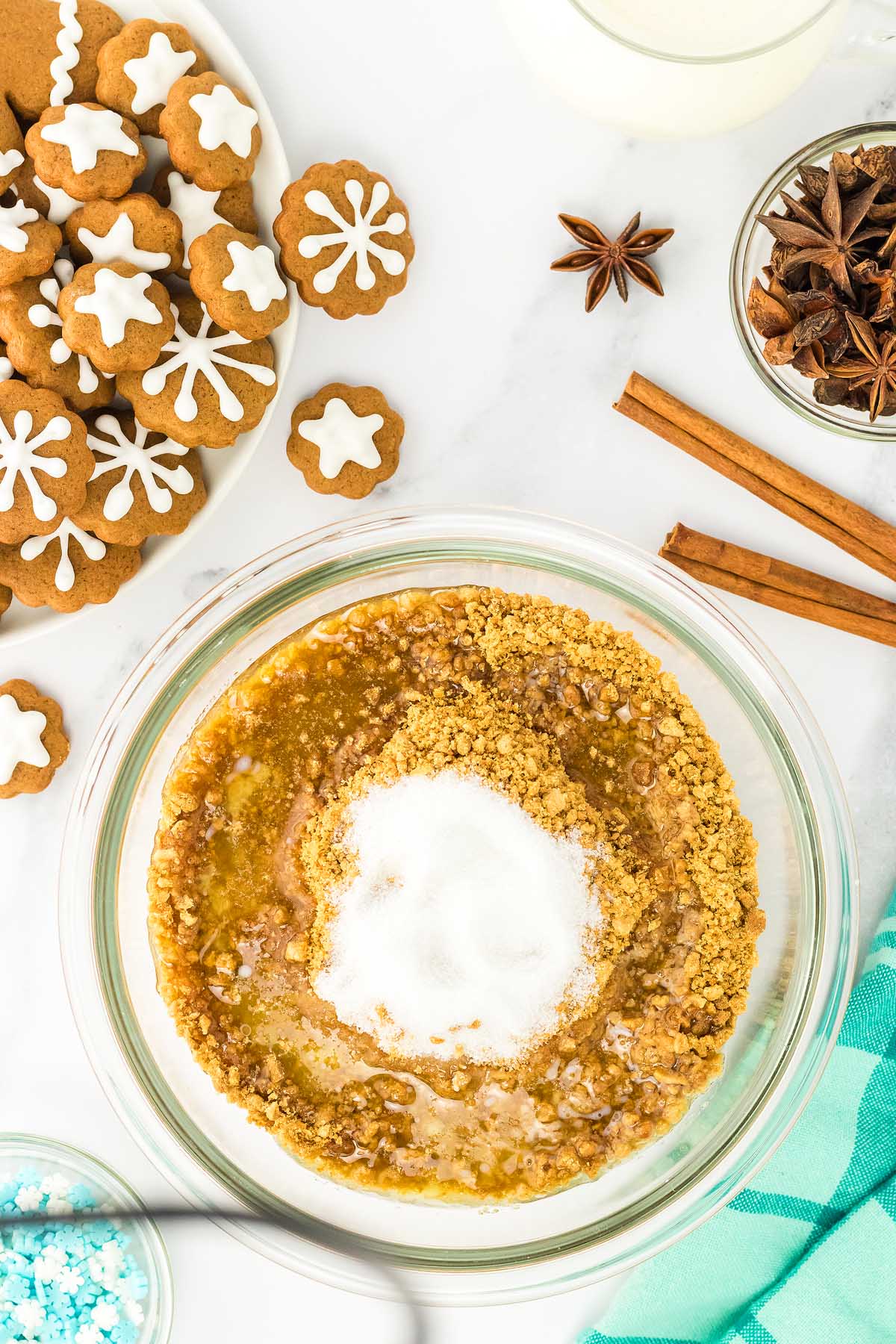 A bowl of crushed graham crackers, sugar, and melted butter sits beside decorated gingerbread cookies and spices, ready to create festive gingerbread cheesecake jars.