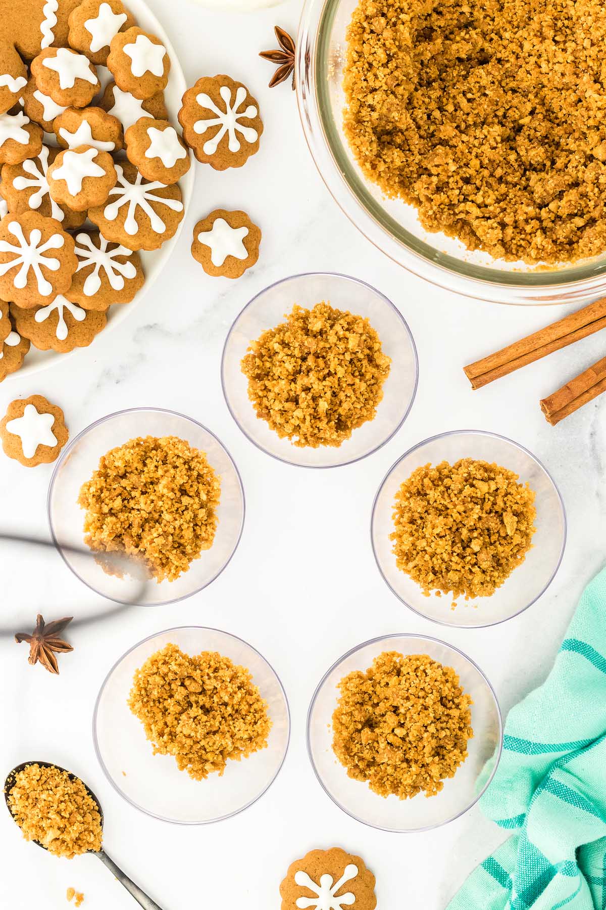Bowls of crumbled cookie crust, gingerbread cheesecake jars, gingerbread cookies with icing, and baking ingredients on a white surface.