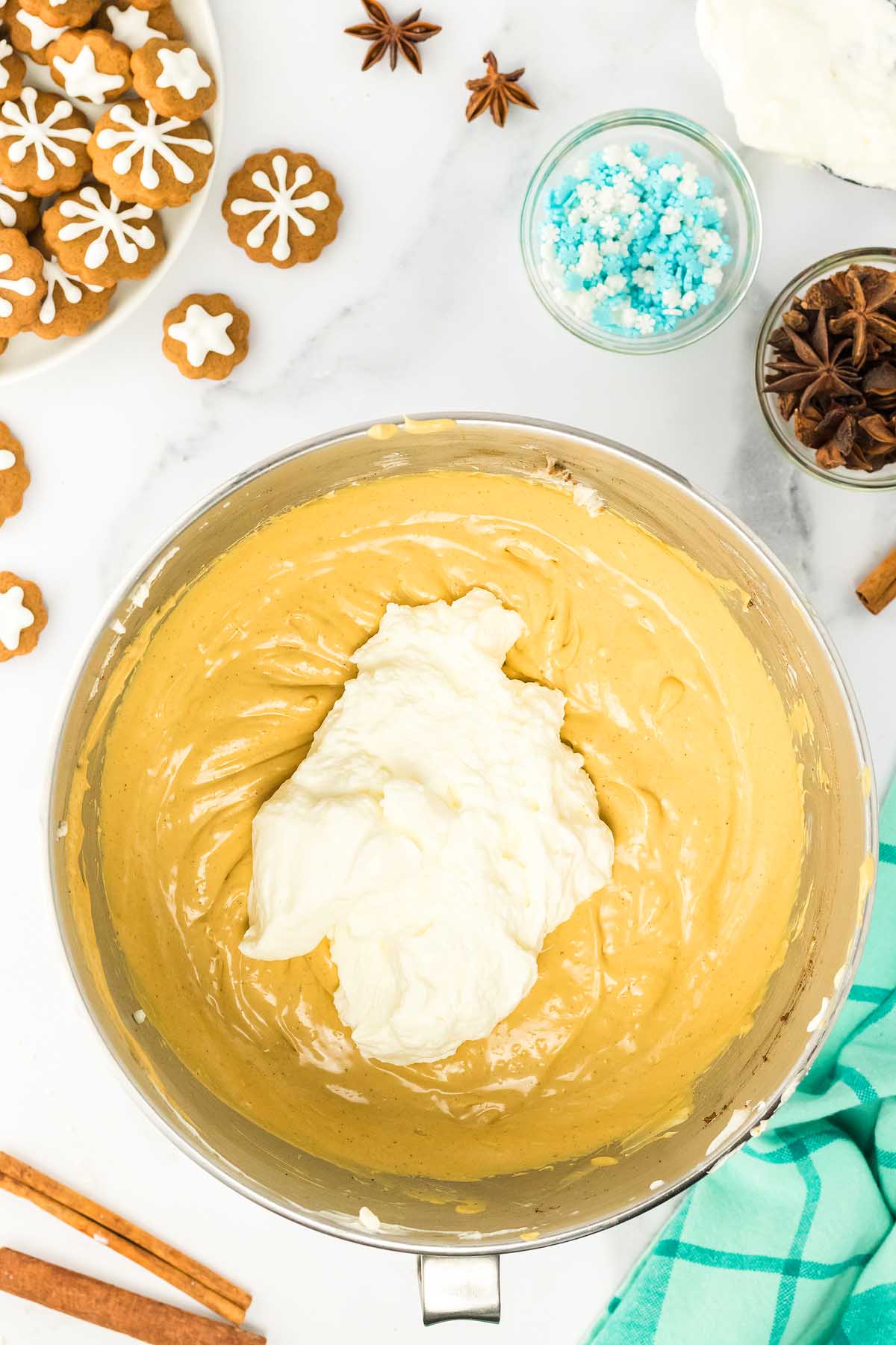 Mixing bowl with whipped cream on top of creamy batter, surrounded by holiday cookies, spices, and festive gingerbread cheesecake jars.
