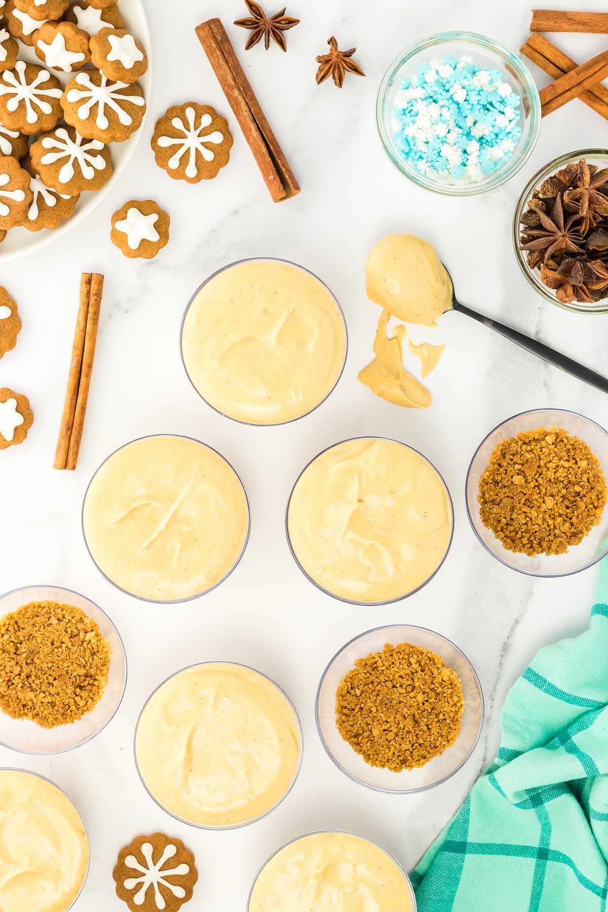 Top view of gingerbread cheesecake jars with creamy filling, crushed cookies, gingerbread cookies, spices, and blue candy on a white surface.