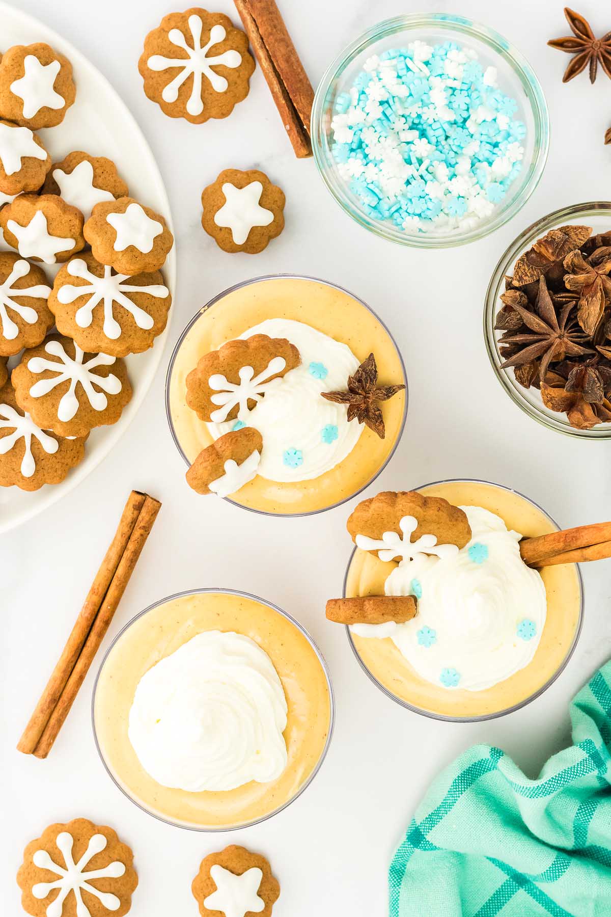 Three glasses of creamy gingerbread cheesecake jars topped with whipped cream, gingerbread cookies, and cinnamon sticks, surrounded by more cookies.