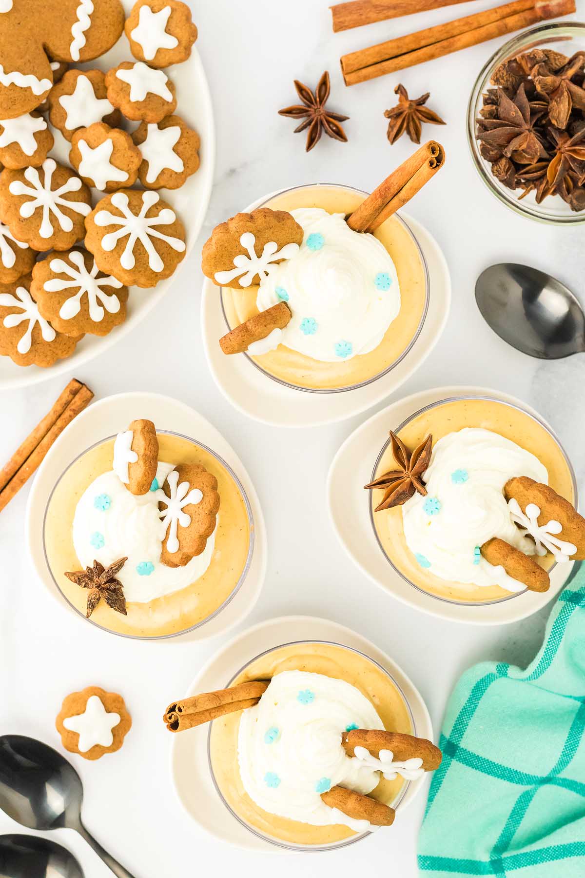 Four gingerbread cheesecake jars topped with whipped cream, gingerbread cookies, cinnamon sticks, and blue sprinkles for a festive creamy dessert.