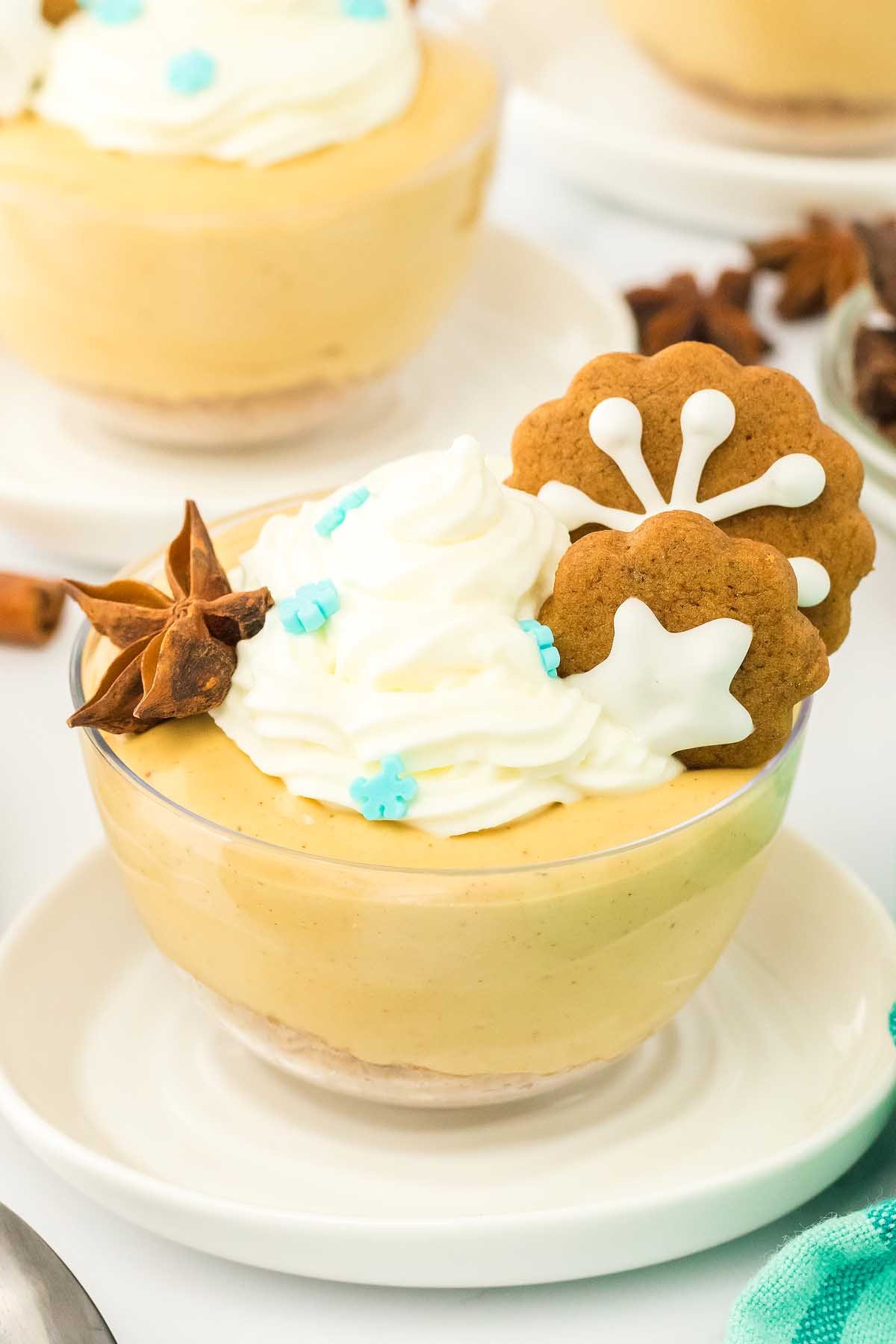 A glass dish of creamy gingerbread cheesecake topped with whipped cream, two iced cookies, and blue sprinkles—perfect for festive cheesecake jars.