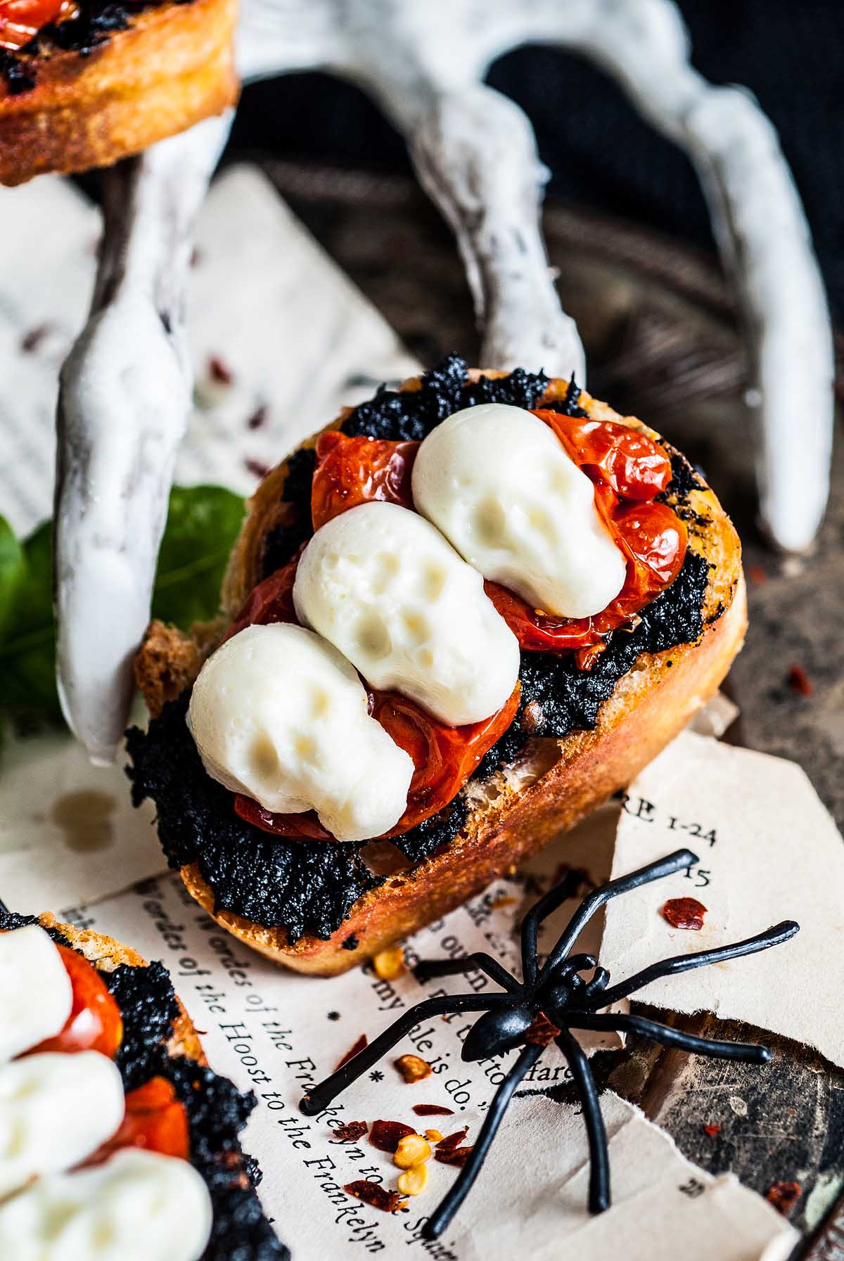 A spooky Halloween bruschetta appetizer featuring mozzarella skulls, black spread, tomatoes, and a fake spider—perfect for your Halloween-themed setup.