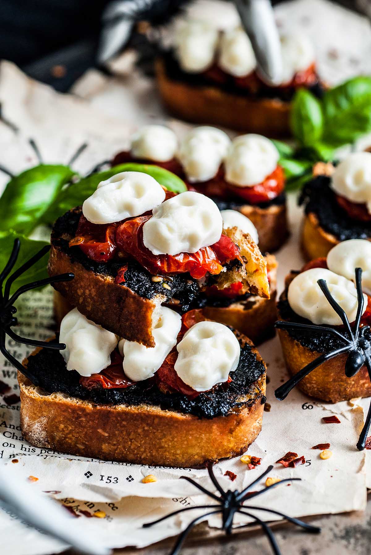 A spooky Halloween bruschetta appetizer featuring toasted bread with a dark spread, tomatoes, and mozzarella shaped like skulls, served alongside creepy fake spiders.