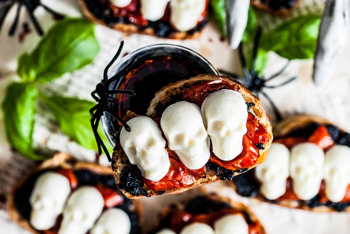 A spooky appetizer featuring toasted bread topped with roasted tomatoes and decorated with small, white skull-shaped mozzarella cheese. The dish is garnished with green basil leaves, black spider decorations, and a glass of dark red liquid in the background.