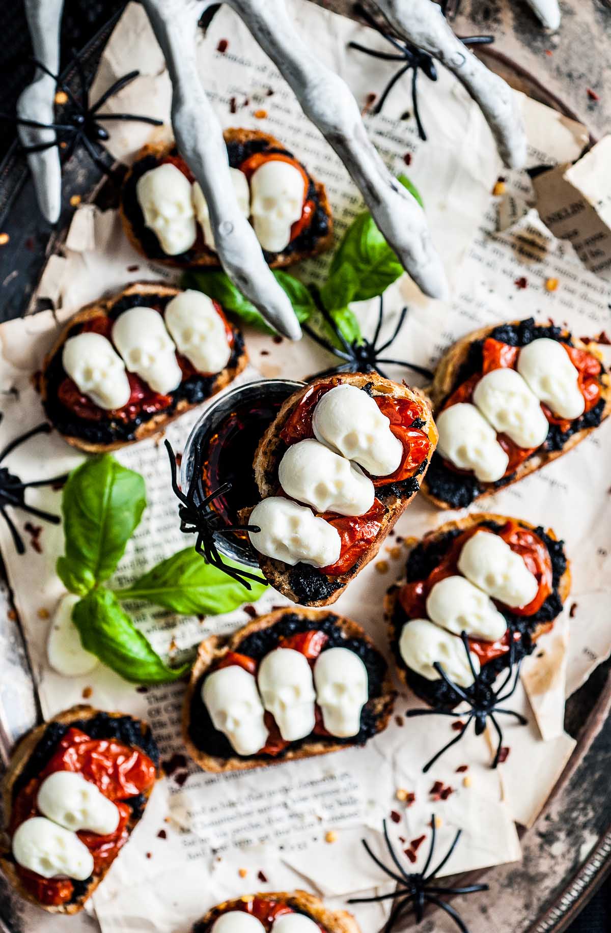 Halloween bruschetta appetizer topped with mozzarella skulls, tomato, fake spiders, and a spooky white skeleton hand—a perfect Halloween appetizer for your festive spread.