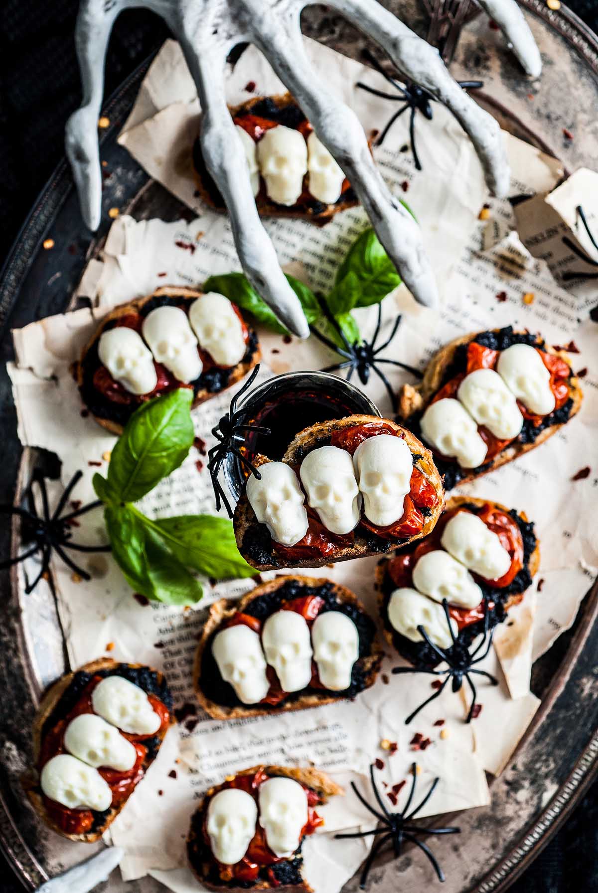 A spooky Halloween bruschetta appetizer topped with white skull shapes, fake spiders, and a creepy skeleton hand decoration—perfect for your Halloween gathering.