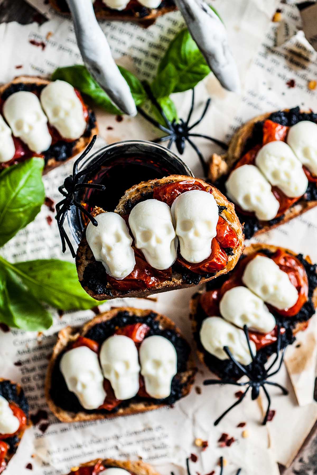 A spooky Halloween bruschetta appetizer featuring toasted bread with tomato topping, mini white skulls, plastic spiders, and basil leaves served on paper.