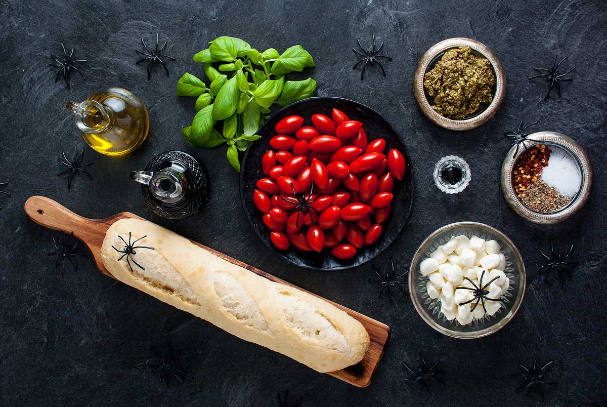 Caprese ingredients, baguette, and fake spiders come together on a dark table for a spooky Halloween bruschetta appetizer setup.