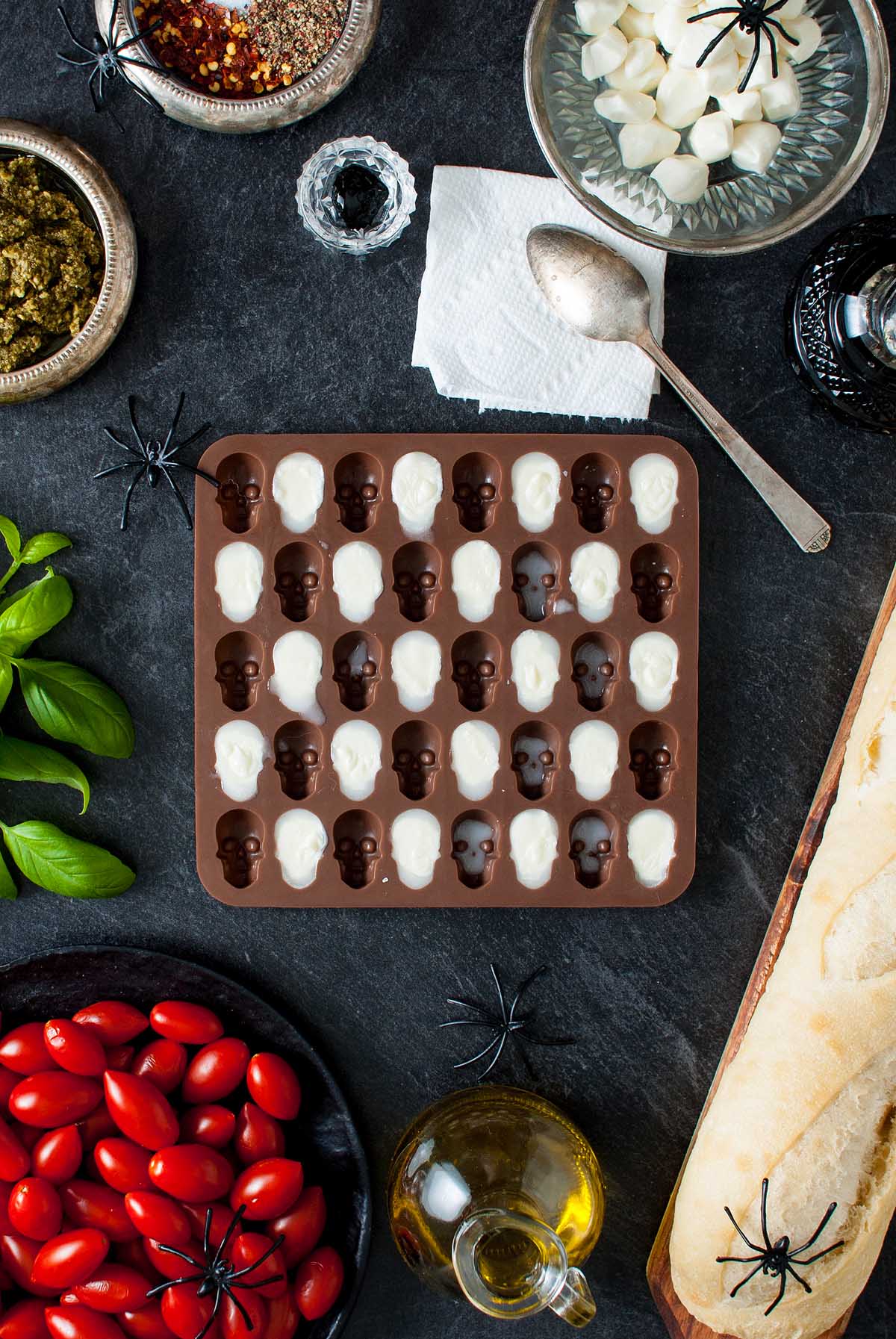 A tray of chocolate and white skull candies is surrounded by Halloween-themed food, spooky plastic spiders, and a festive Halloween bruschetta appetizer.