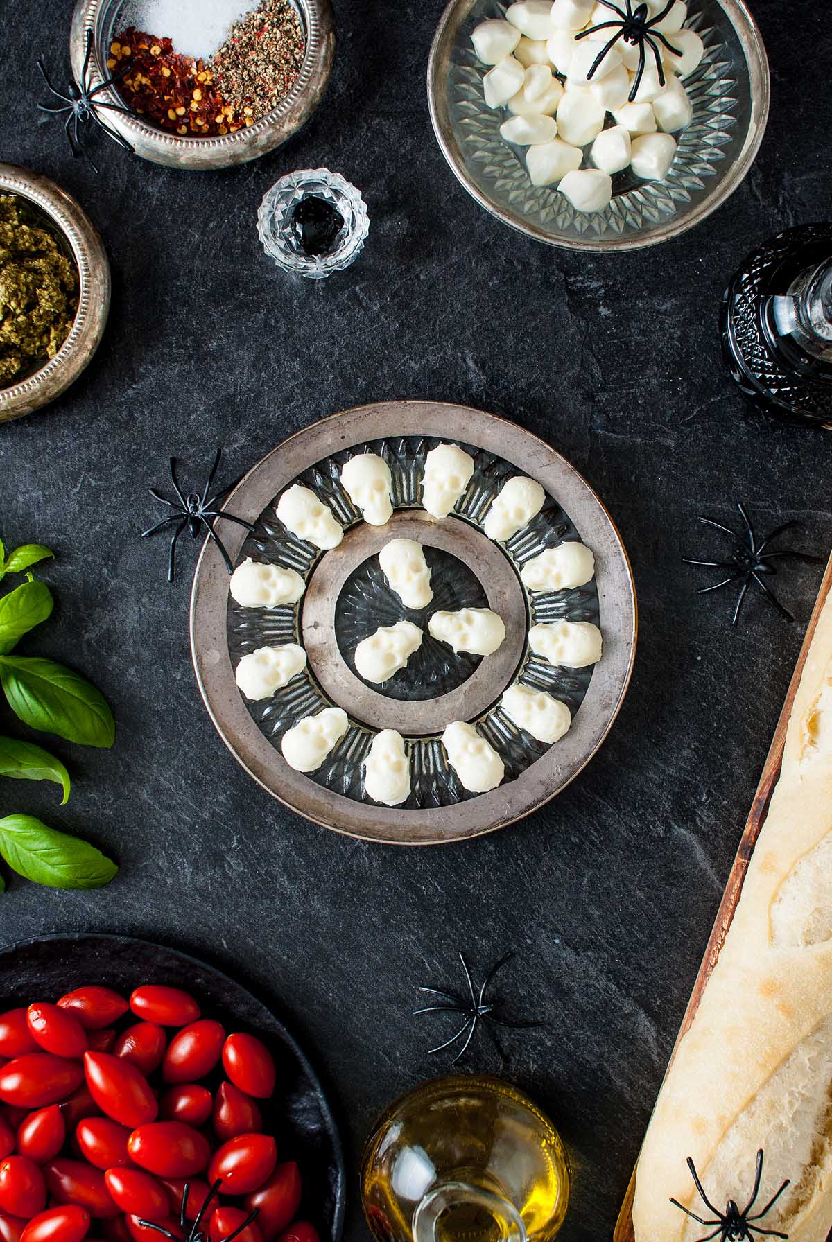 A plate of ghost-shaped mozzarella bites makes a fun Halloween appetizer, surrounded by festive props, basil, tomatoes, and bread—perfect for a spooky Halloween bruschetta appetizer spread.