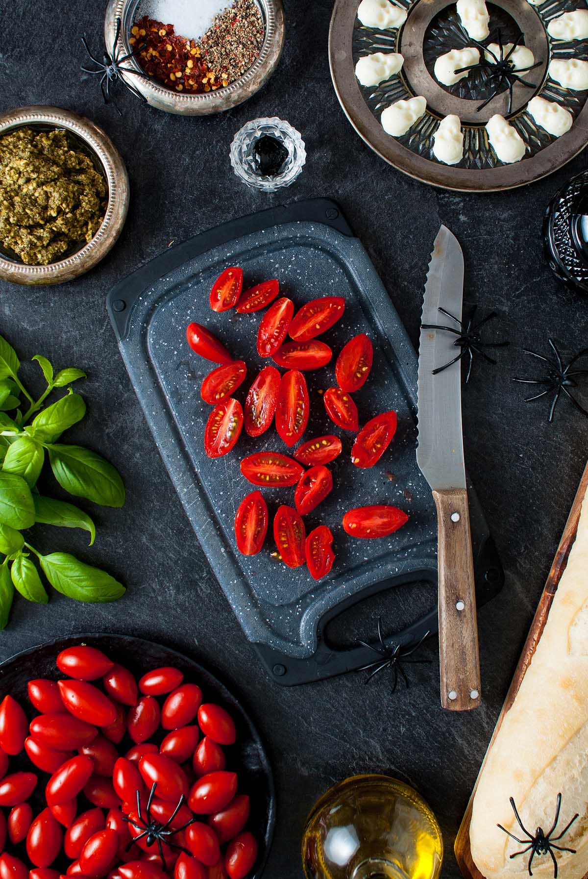 Sliced grape tomatoes on a cutting board surrounded by ingredients and fake spiders on a dark surface, perfect for a spooky Halloween bruschetta appetizer.