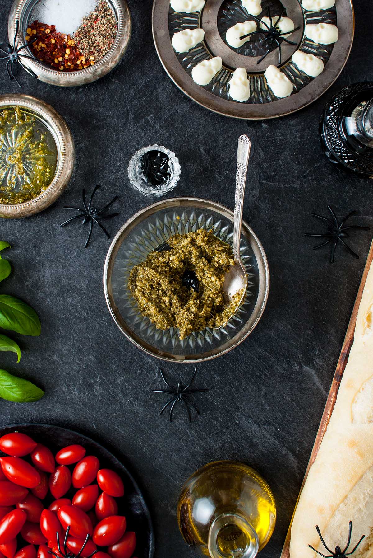 Flat lay of a Halloween bruschetta appetizer: pesto in a bowl, tomatoes, bread, oil, herbs, and festive Halloween spider decorations on a dark table.