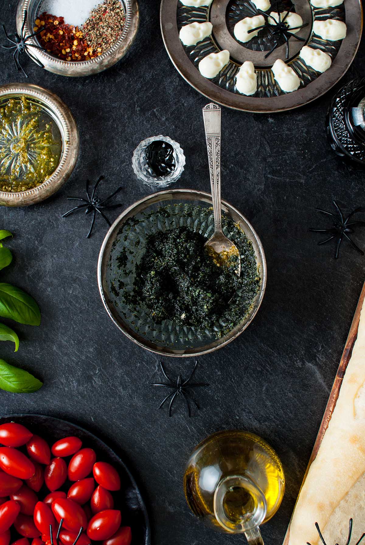 A bowl of black pesto with a spoon, surrounded by tomatoes, basil, oil, spices, and Halloween decorations—the perfect Halloween bruschetta appetizer for your spooky celebration.