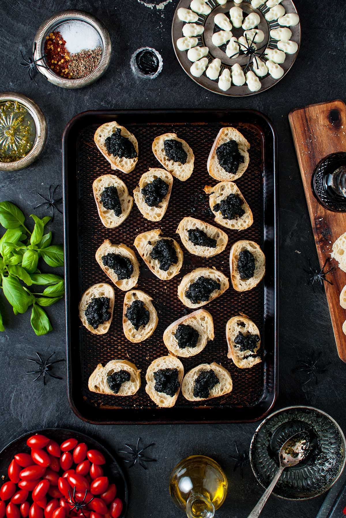 Tray of bread slices with black spread, surrounded by tomatoes, basil, cheese, oil, and festive Halloween decorations—a perfect Halloween bruschetta appetizer for your spooky gathering.