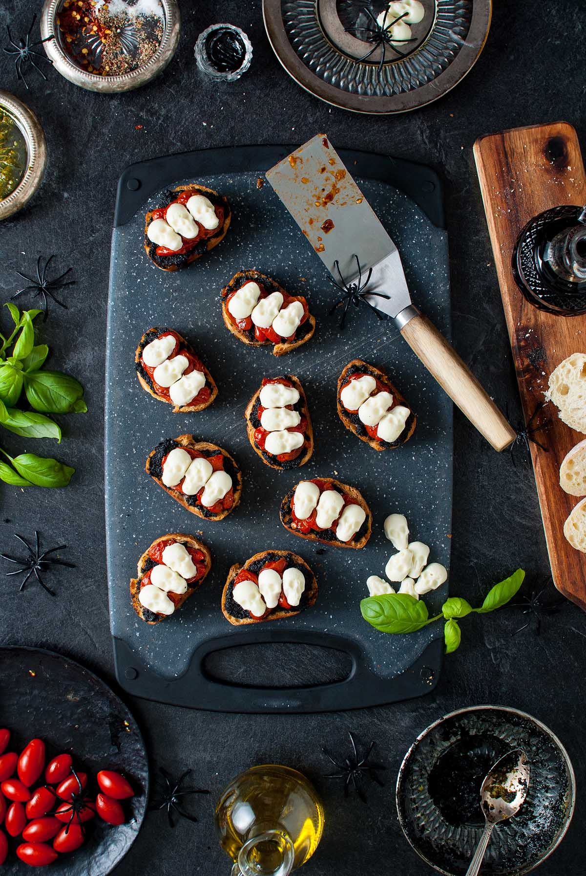Bruschetta with mozzarella and tomato on a cutting board, surrounded by basil, plates, and Halloween decor—create the perfect Halloween bruschetta appetizer for your spooky gathering.