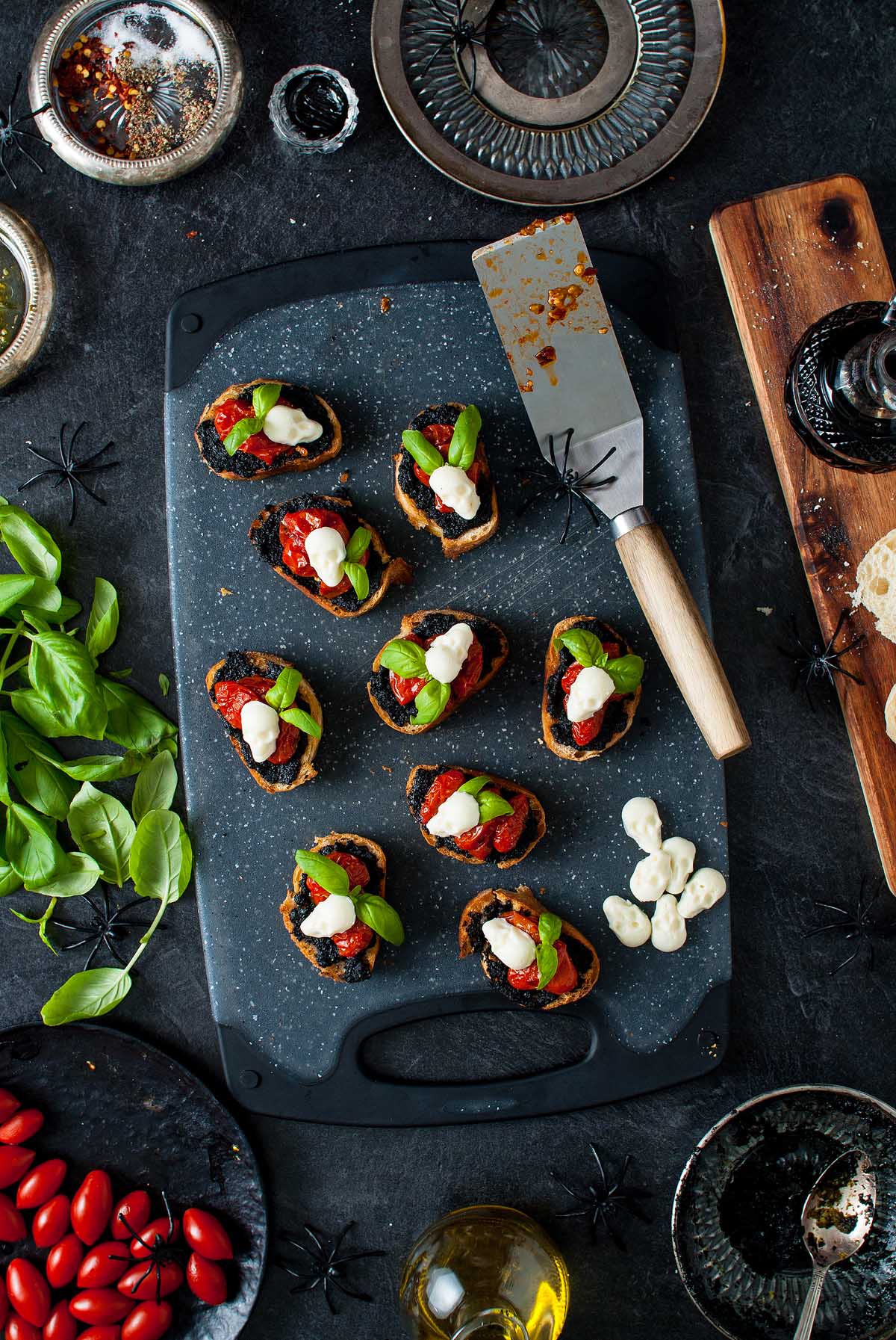Nine Halloween bruschetta appetizers with basil and mozzarella are artfully arranged on a black cutting board, surrounded by spooky decor for a festive Halloween appetizer display.