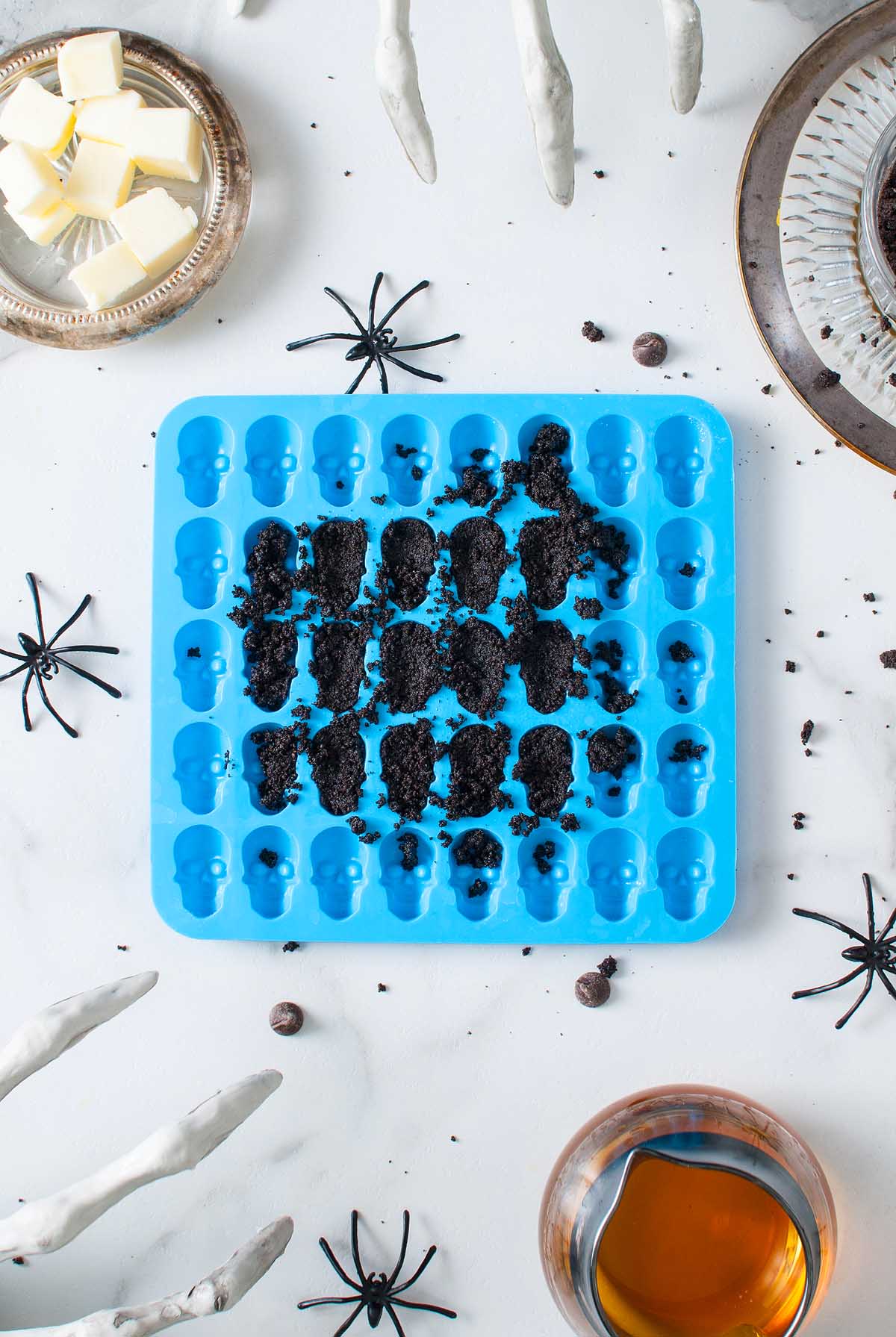 Blue skull-shaped silicone mold filled with crushed cookies, surrounded by fake spiders and Halloween decorations—an eerie Boozy Halloween Pot de Creme Dessert that's perfect for your next spooky Halloween dessert table.
