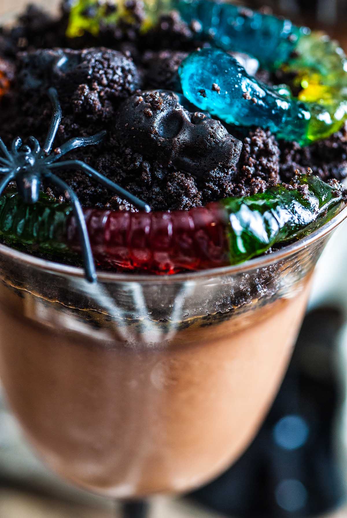 Chocolate pudding topped with crushed cookies, gummy worms, and a plastic spider for a spooky Boozy Halloween Pot de Creme Dessert that's perfect as a fun Halloween dessert.