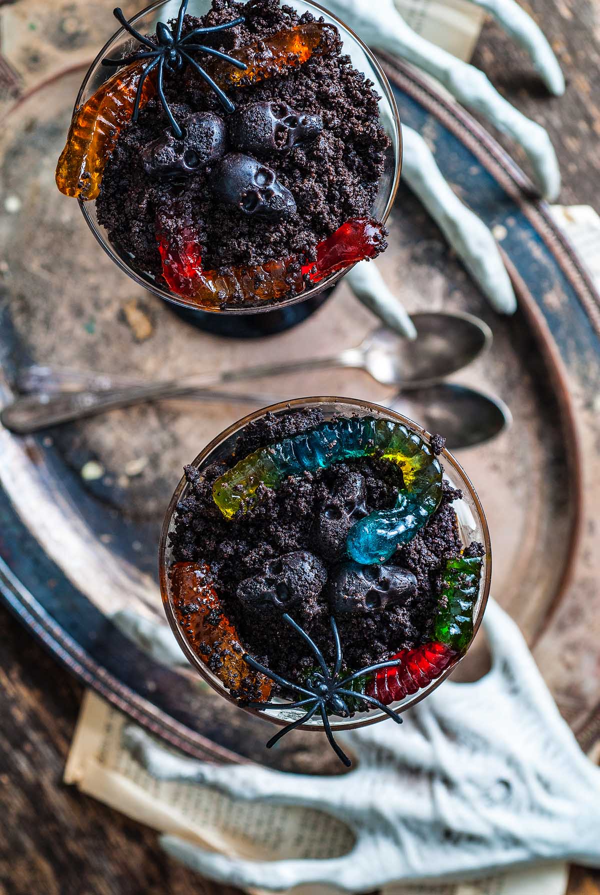 Two chocolate dirt cups topped with gummy worms and fake spiders, set on a tray with skeleton hands and spoons—this Boozy Halloween Pot de Creme Dessert makes the ultimate spooky Halloween dessert.