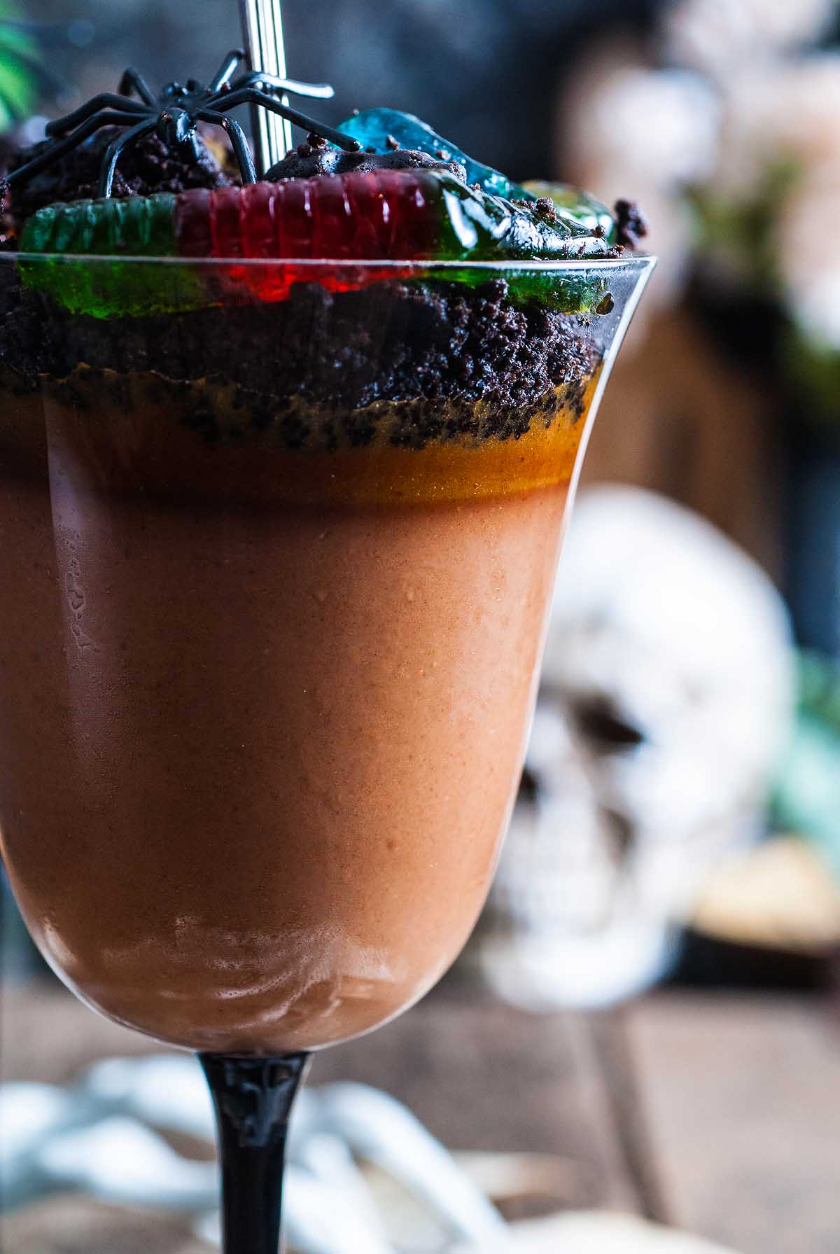 Chocolate pudding in a glass, topped with cookie crumbs and gummy worms, makes for a spooky Halloween dessert—complete with a skull decoration in the background.