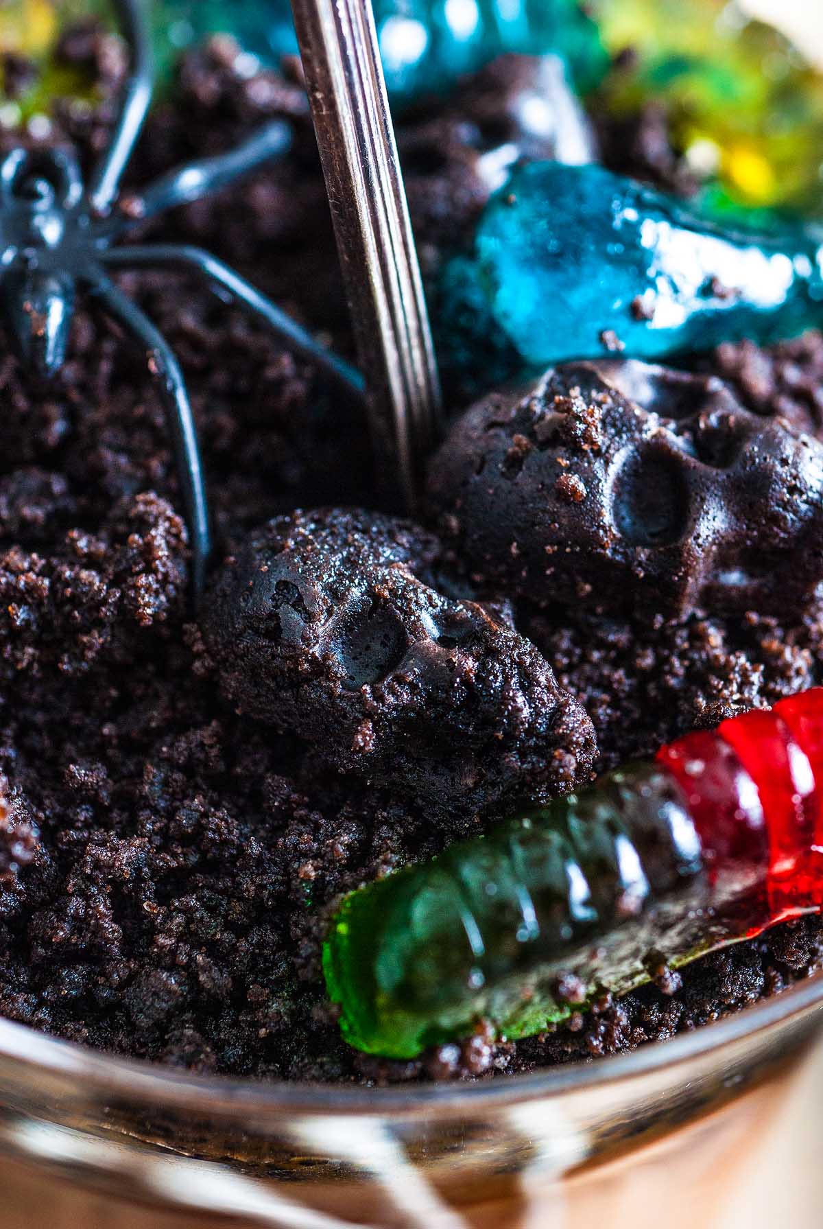 Chocolate cookie crumbs with gummy worms, a plastic spider, and candy skulls create a spooky Halloween dessert scene perfect for your Boozy Halloween Pot de Creme Dessert.