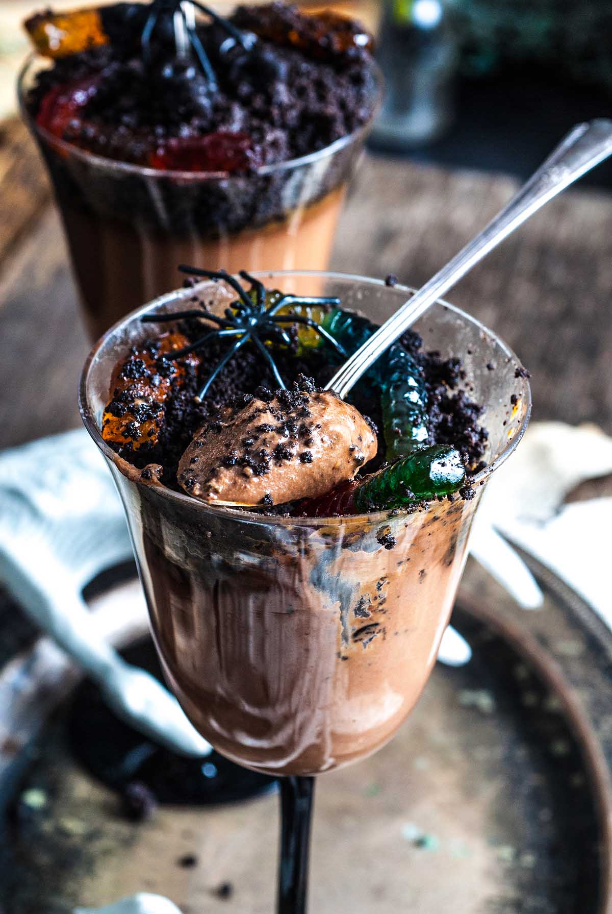 Halloween dessert chocolate mousse in a glass, topped with cookie crumbs, gummy worms, and a plastic spider, with a spoon tucked inside for the ultimate spooky treat.