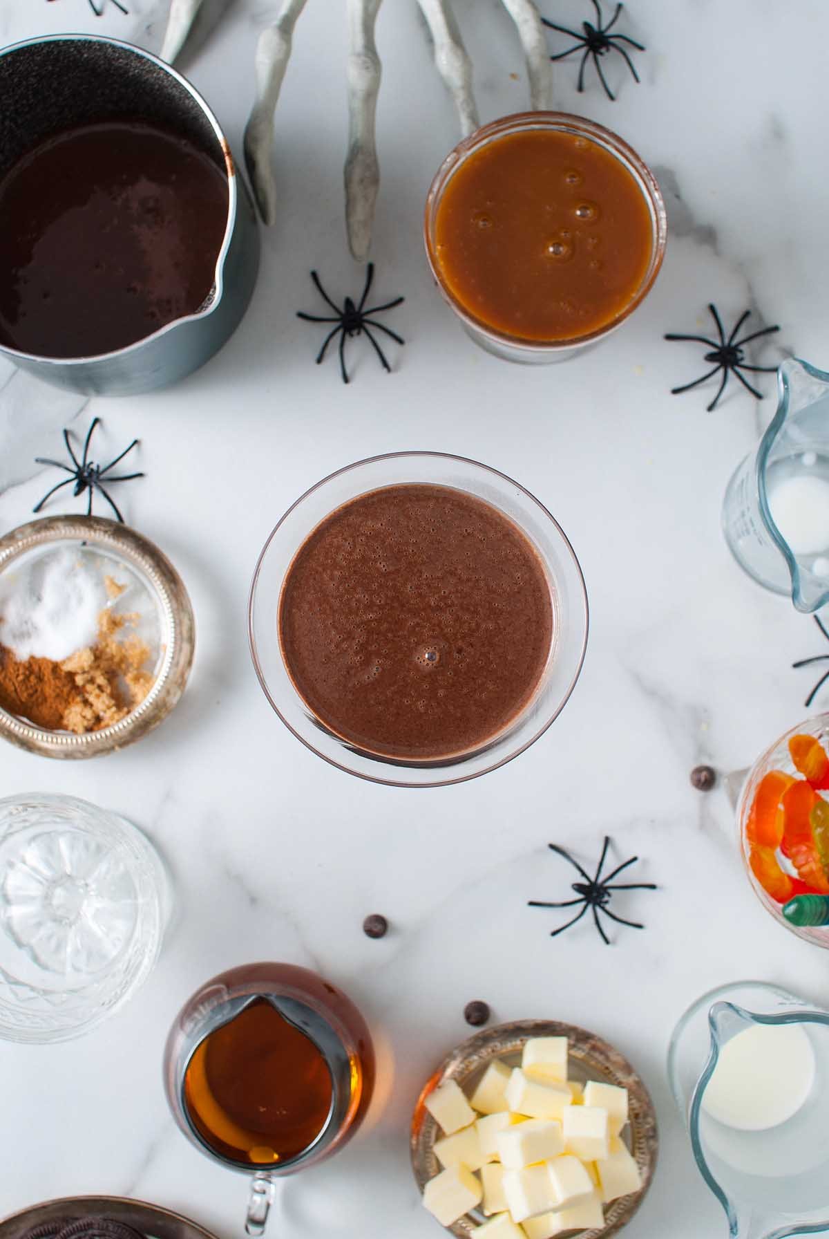 Overhead view of a Boozy Halloween Pot de Creme Dessert, chocolate drink, caramel, and baking ingredients on a marble table with fake spiders and a skeleton hand, perfect for a spooky Halloween dessert spread.