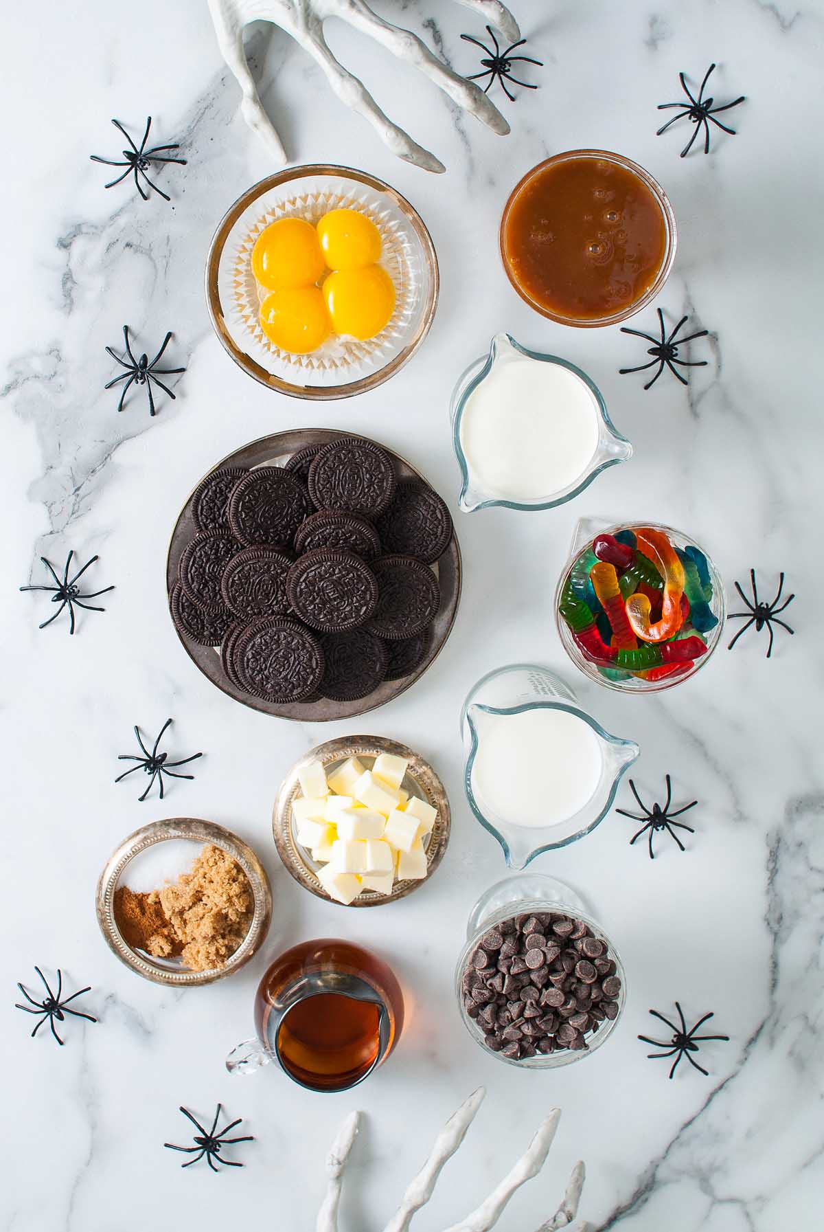 Assorted Halloween dessert ingredients with cookies, chocolate chips, gummy worms, and candy eyeballs on marble with fake spiders—perfect for crafting a festive Boozy Halloween Pot de Creme Dessert.