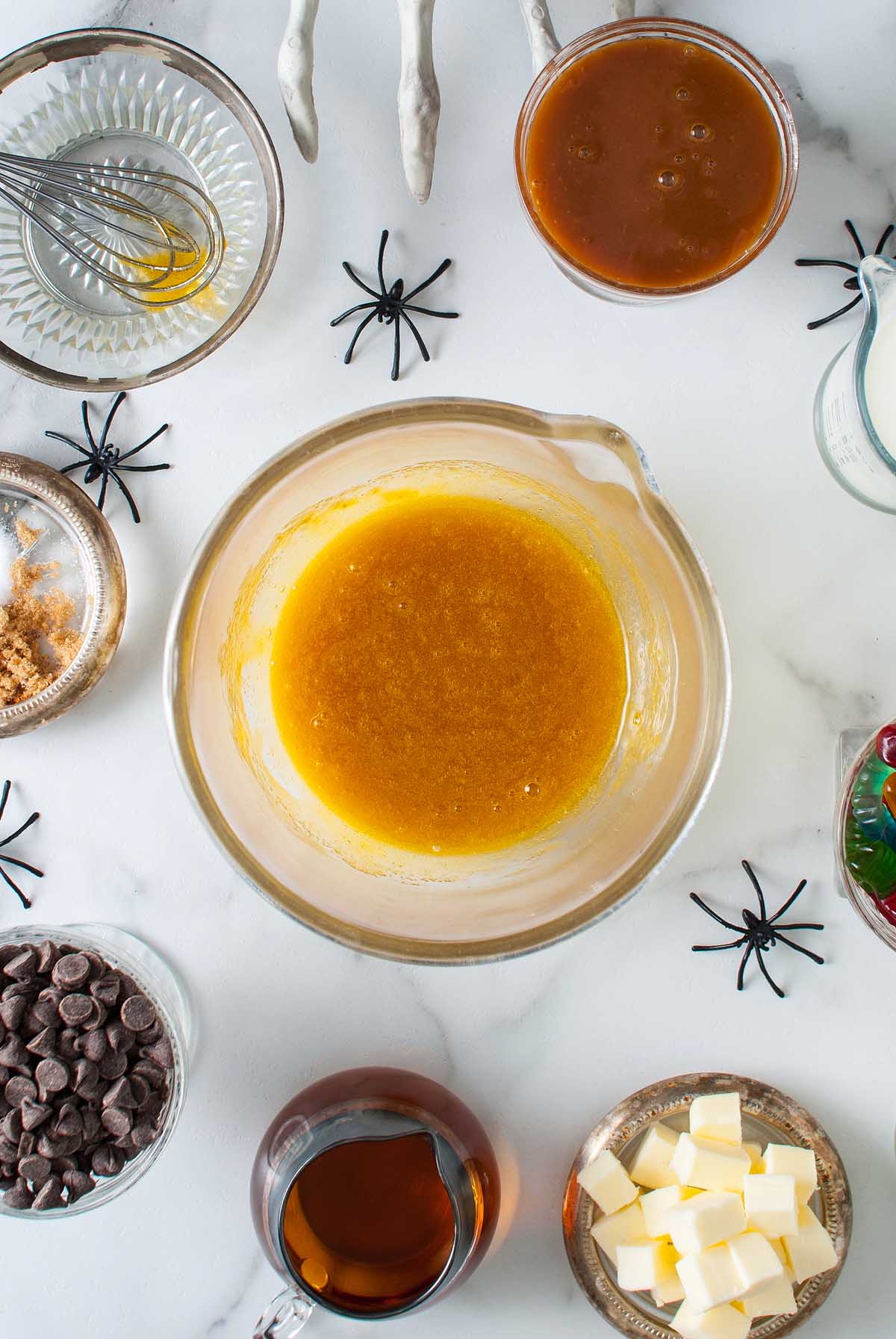 Overhead view of baking ingredients in bowls, with chocolate chips, butter, and spider decorations on a white surface—perfect for creating a Boozy Halloween Pot de Creme Dessert or any festive Halloween dessert.