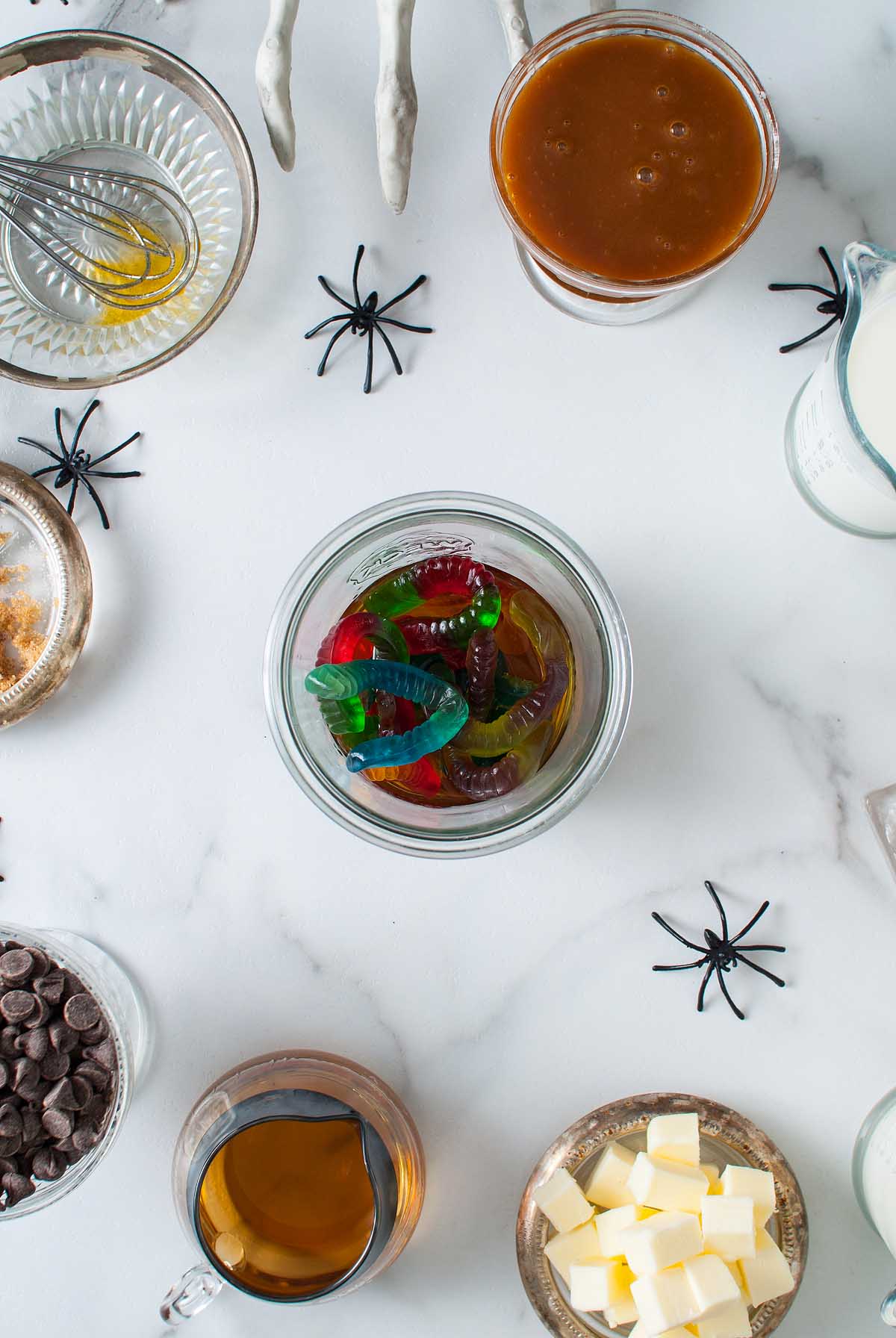 A jar of gummy worms surrounded by chocolate chips, butter cubes, caramel, and fake spiders sits on a white surface—perfect as a playful Halloween dessert.