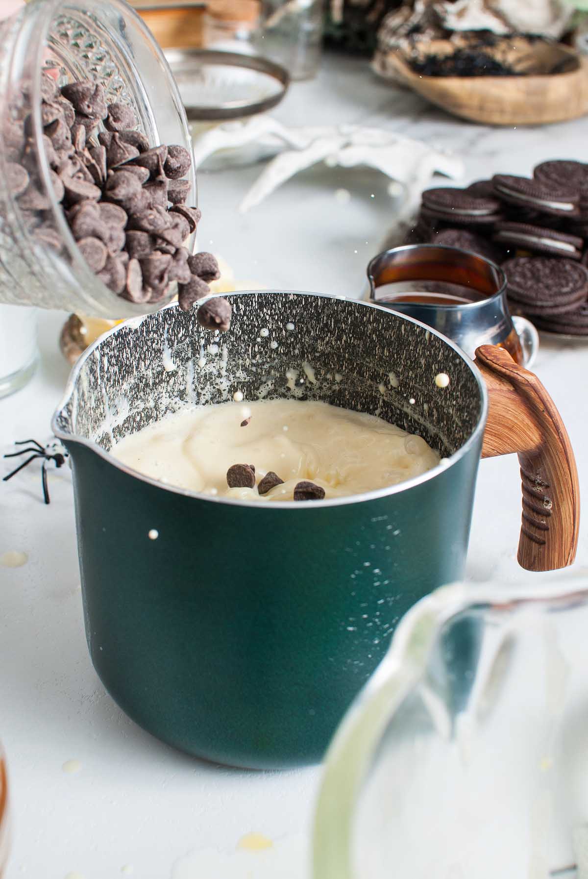 Chocolate chips are being poured into a saucepan of creamy mixture on a kitchen counter, preparing a delicious Boozy Halloween Pot de Creme Dessert.