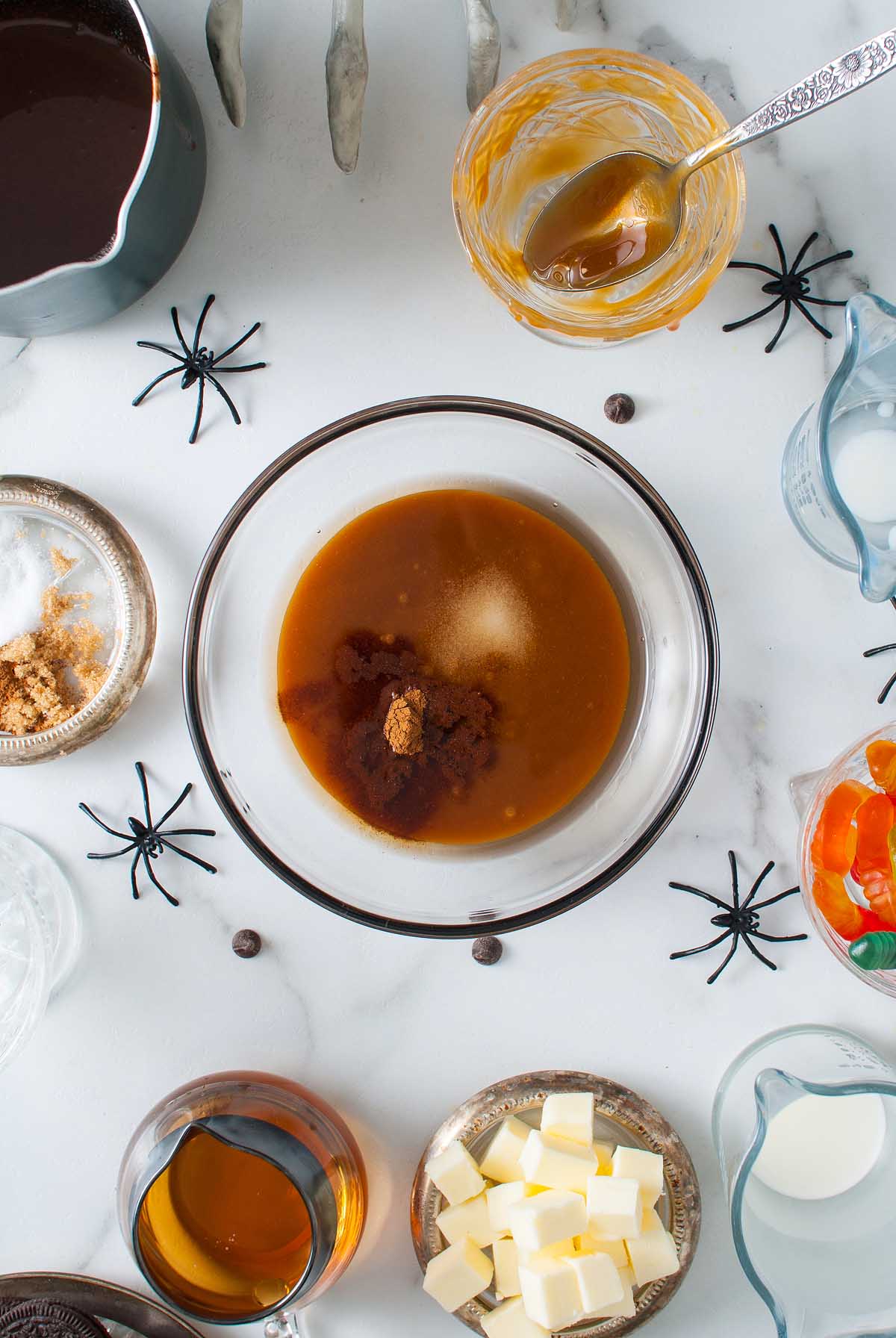Glass bowl with brown liquid and spices on a white table, surrounded by candy, butter, and fake black spiders—this Boozy Halloween Pot de Creme Dessert is the ultimate Halloween dessert centerpiece.