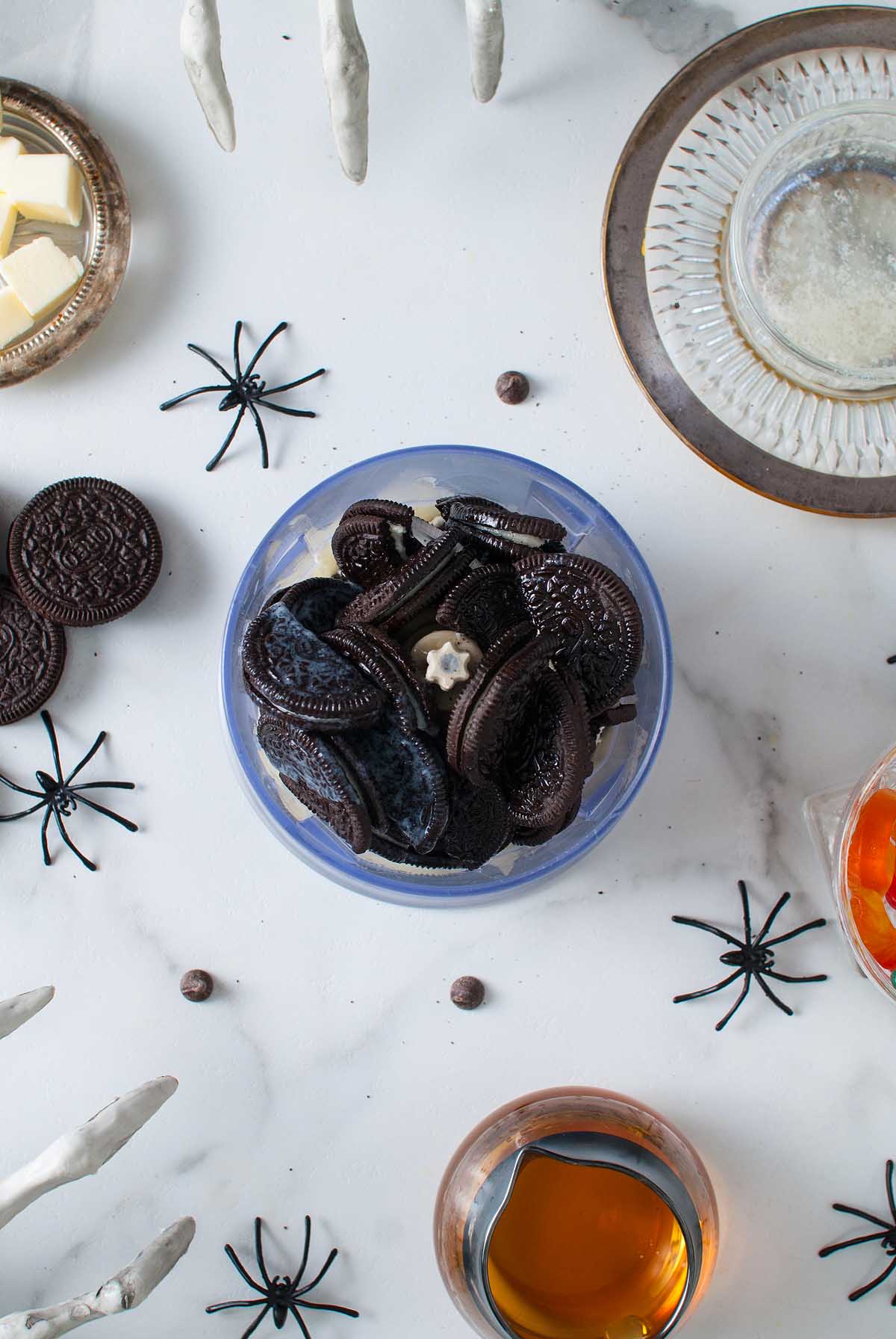 A bowl of chocolate sandwich cookies, perfect as a Halloween dessert, is surrounded by fake spiders and drinks on a white surface.