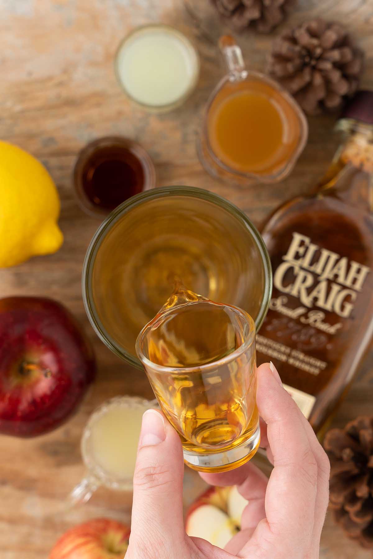 A hand pours whiskey from a shot glass into a glass, surrounded by apple, lemon, and cocktail ingredients—perfect for crafting a Maple Apple Whiskey Sour.