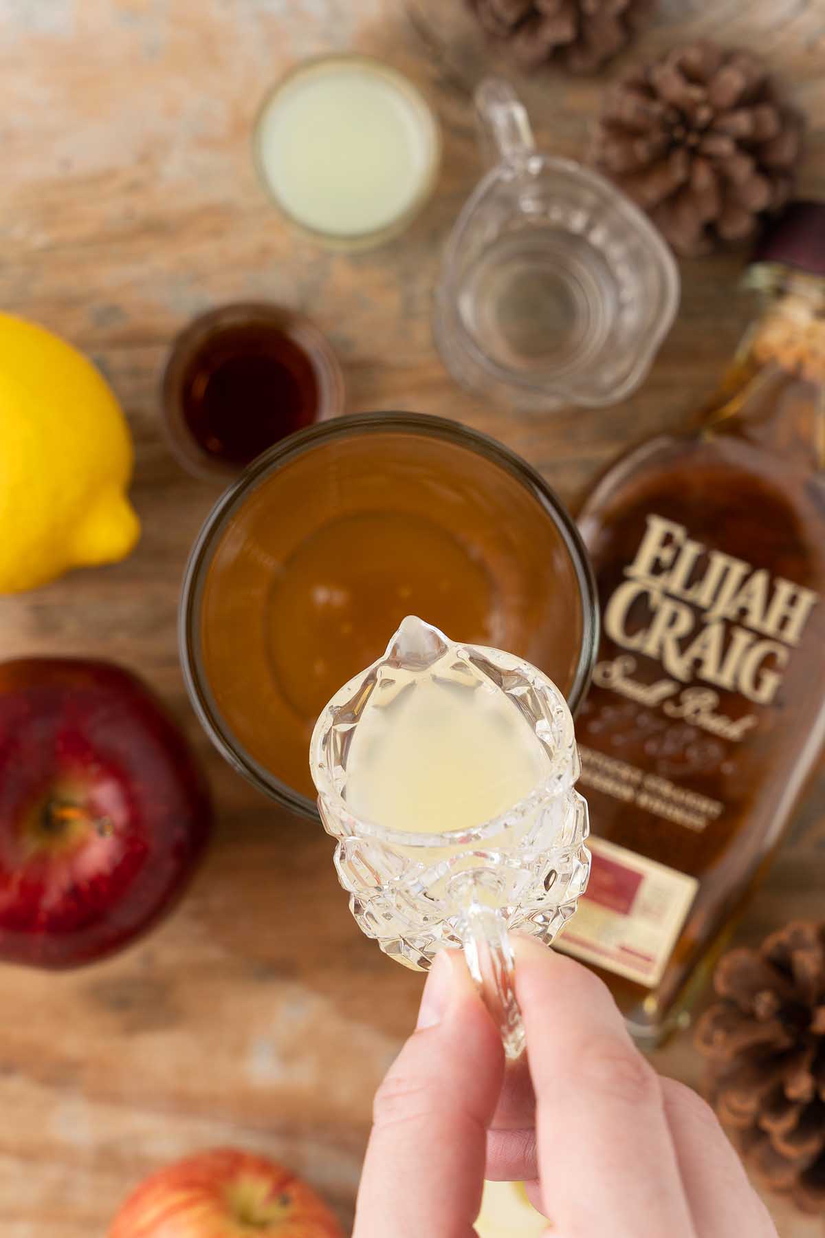 A hand pours liquid from a small glass into a drink, surrounded by whiskey, apple, lemon, and ingredients—perfect for crafting a Maple Apple Whiskey Sour.