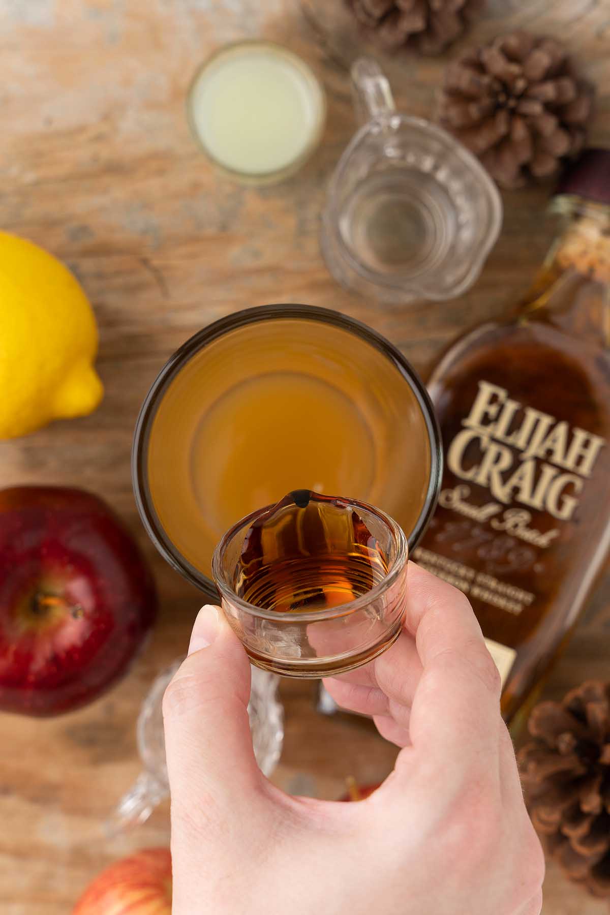 A hand holds a shot glass above a cocktail, surrounded by whiskey, an apple, a lemon, and mixing ingredients—perfect for crafting a Maple Apple Whiskey Sour.