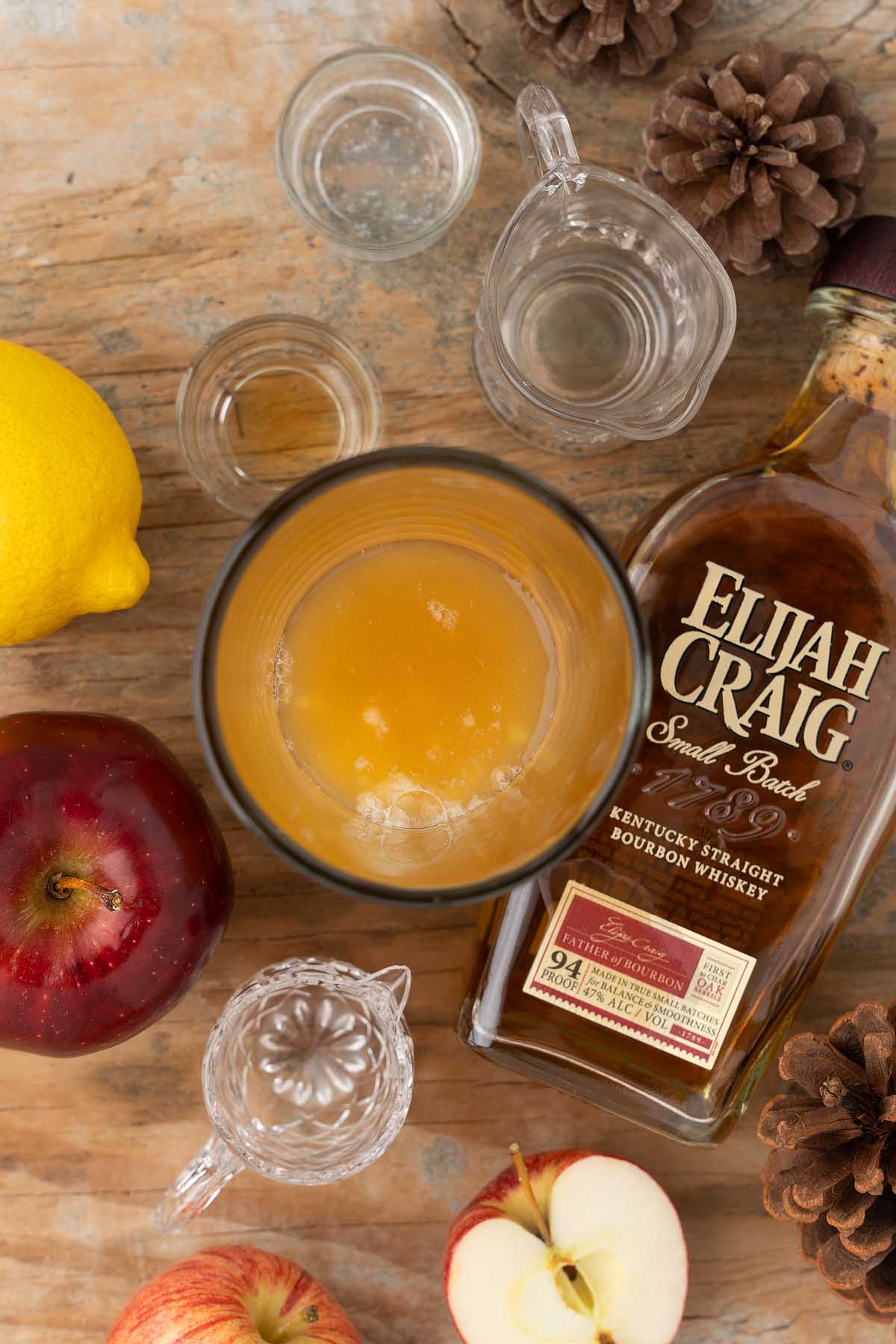 Top view of a Maple Apple Whiskey Sour in a glass, surrounded by a whiskey bottle, apple, lemon, apple slices, and small pitchers on the table.