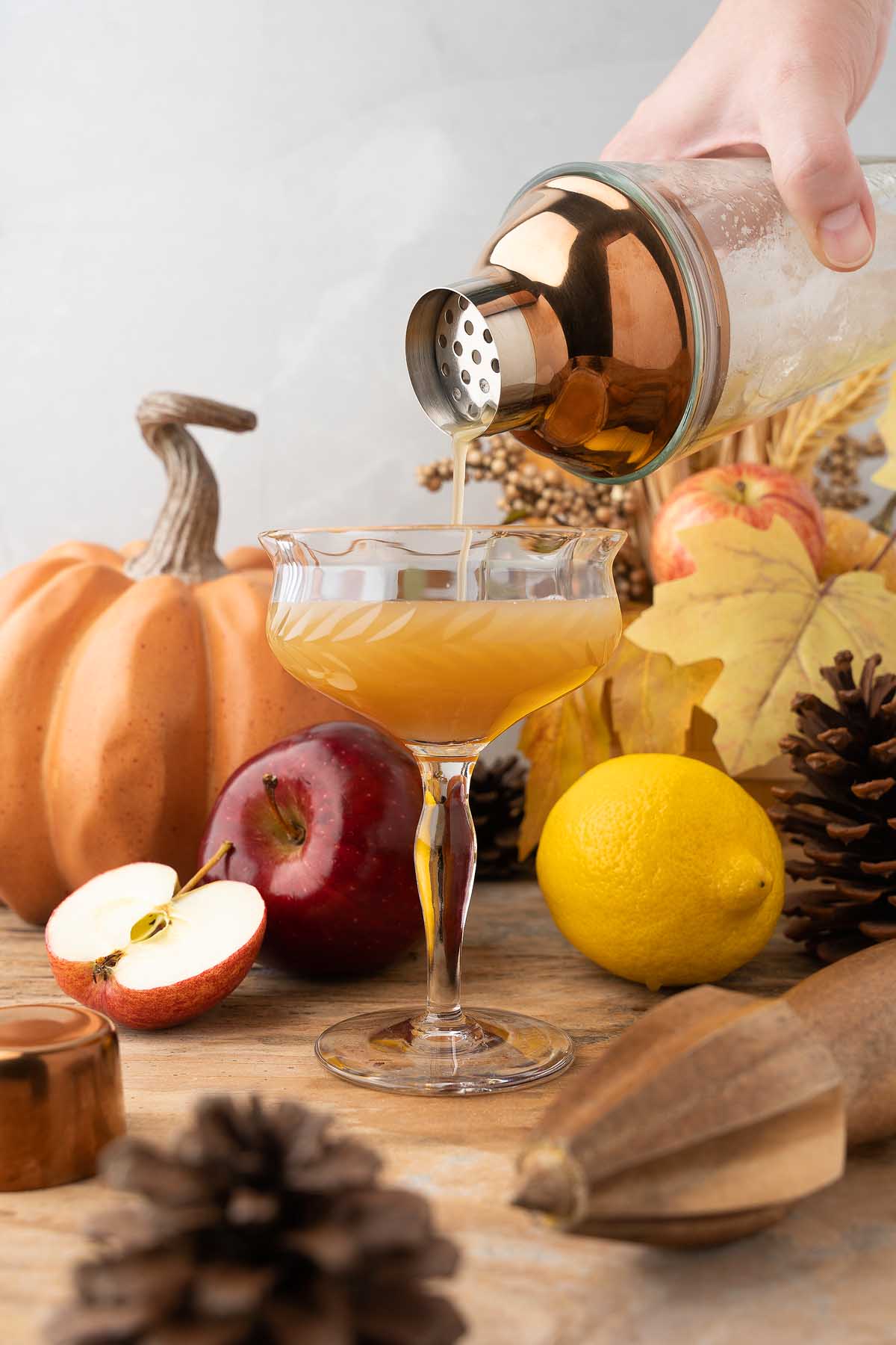 A hand pours a Maple Apple Whiskey Sour into a glass, surrounded by fall decor, pumpkin, apple, lemon, and pinecones.