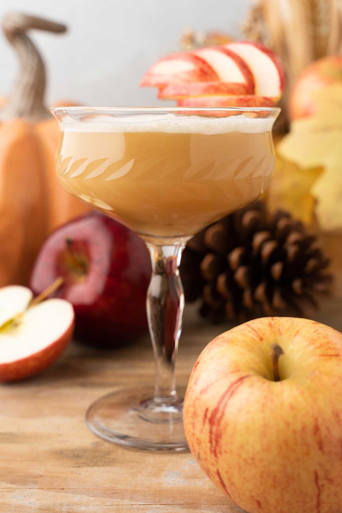 A frothy Maple Apple Whiskey Sour in a coupe glass, garnished with apple slices, surrounded by fresh apples and autumn decor.