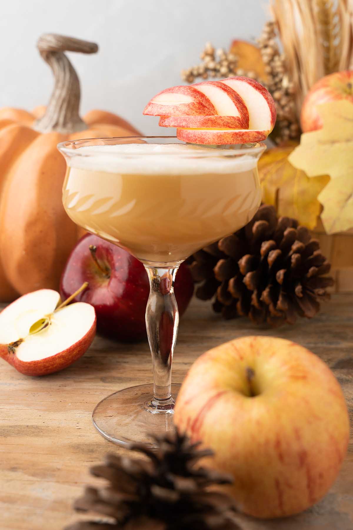 A Maple Apple Whiskey Sour fall cocktail garnished with apple slices, surrounded by pumpkins, apples, pinecones, and autumn leaves.