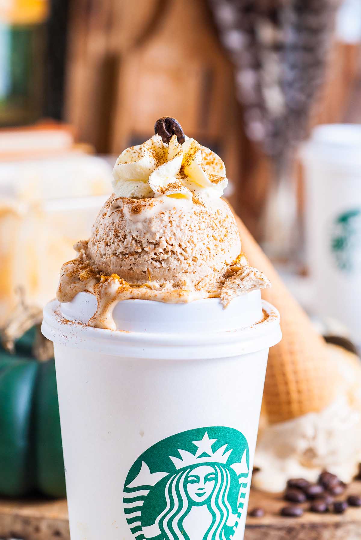 A Starbucks cup topped with a scoop of no churn pumpkin spice latte ice cream, whipped cream, and a coffee bean, surrounded by coffee beans.